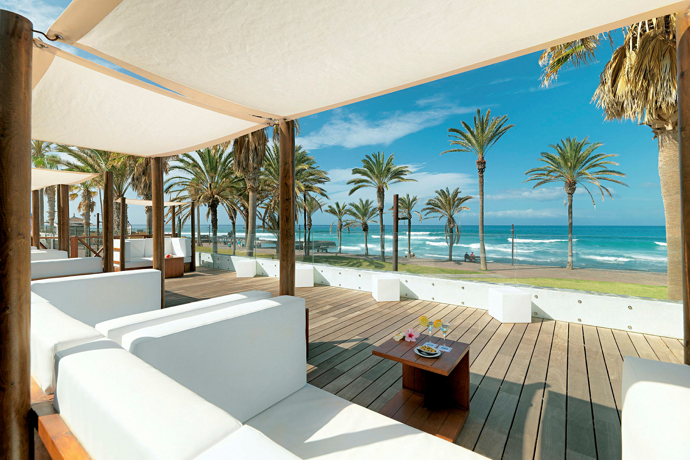 En udendørs lounge med hvide sofaer og parasoller med udsigt over stranden og havet. (AI-genereret tekst)