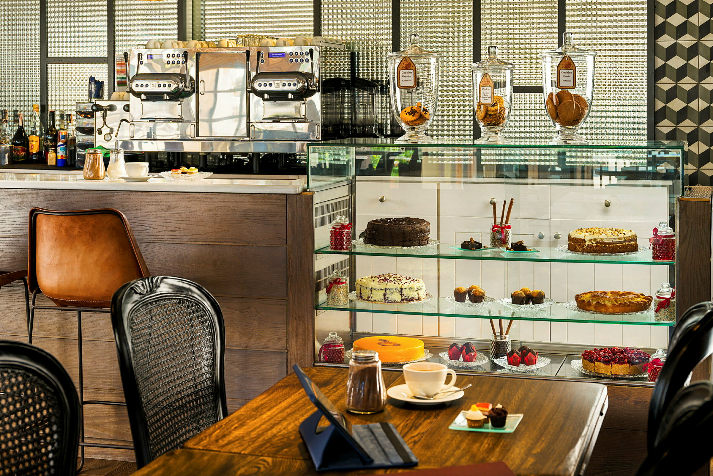 Et billede af en café med kager og bagværk i en glasmontre, en espressomaskine og elegante kaffekopper på en bordflade. (AI-genereret tekst)