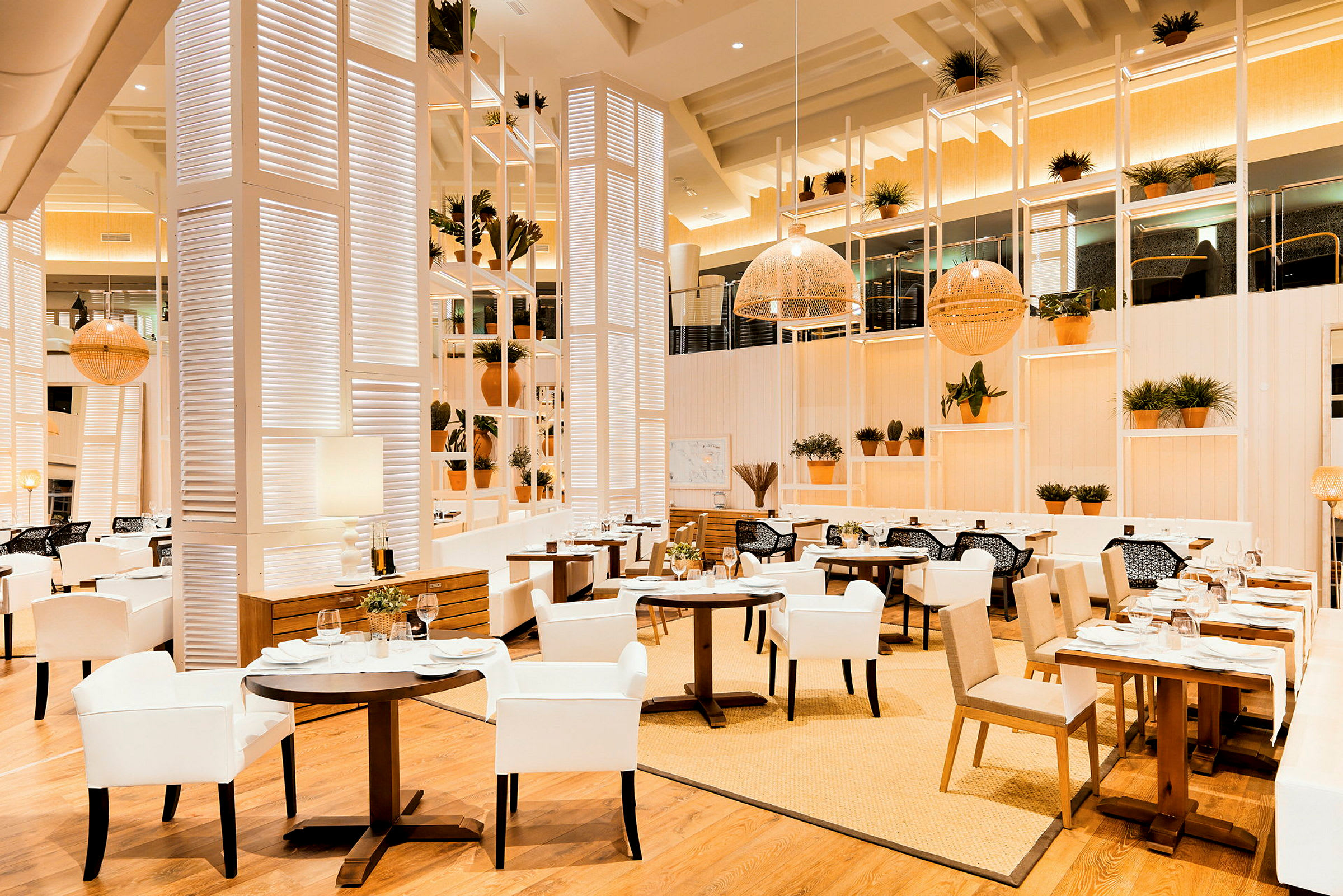 En elegant restaurant med lys indretning, hvide og beige møbler samt træmaterialer. Potteplanter pryder hylderne og bordene. (AI-genereret tekst)