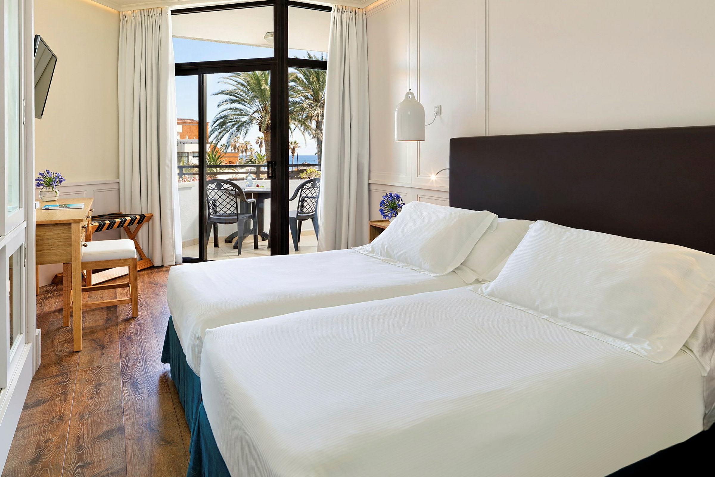 Et lyst hotelværelse med to enkeltsenge, et skrivebord og en balkon med udsigt over havet og palmer. (AI-genereret tekst)