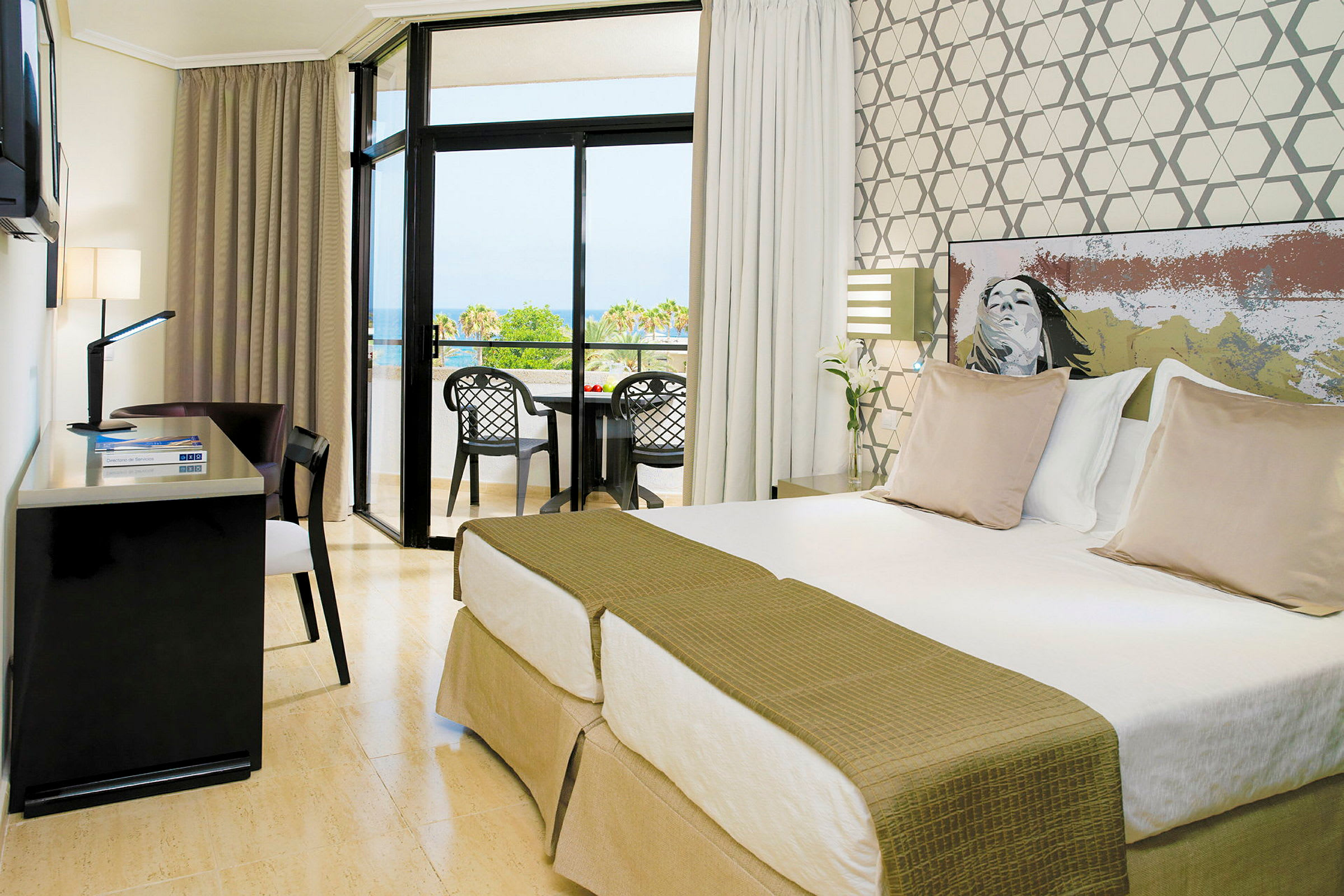Et hotelværelse med to enkeltsenge, et skrivebord og en balkon med udsigt over et grønt område og havet. (AI-genereret tekst)