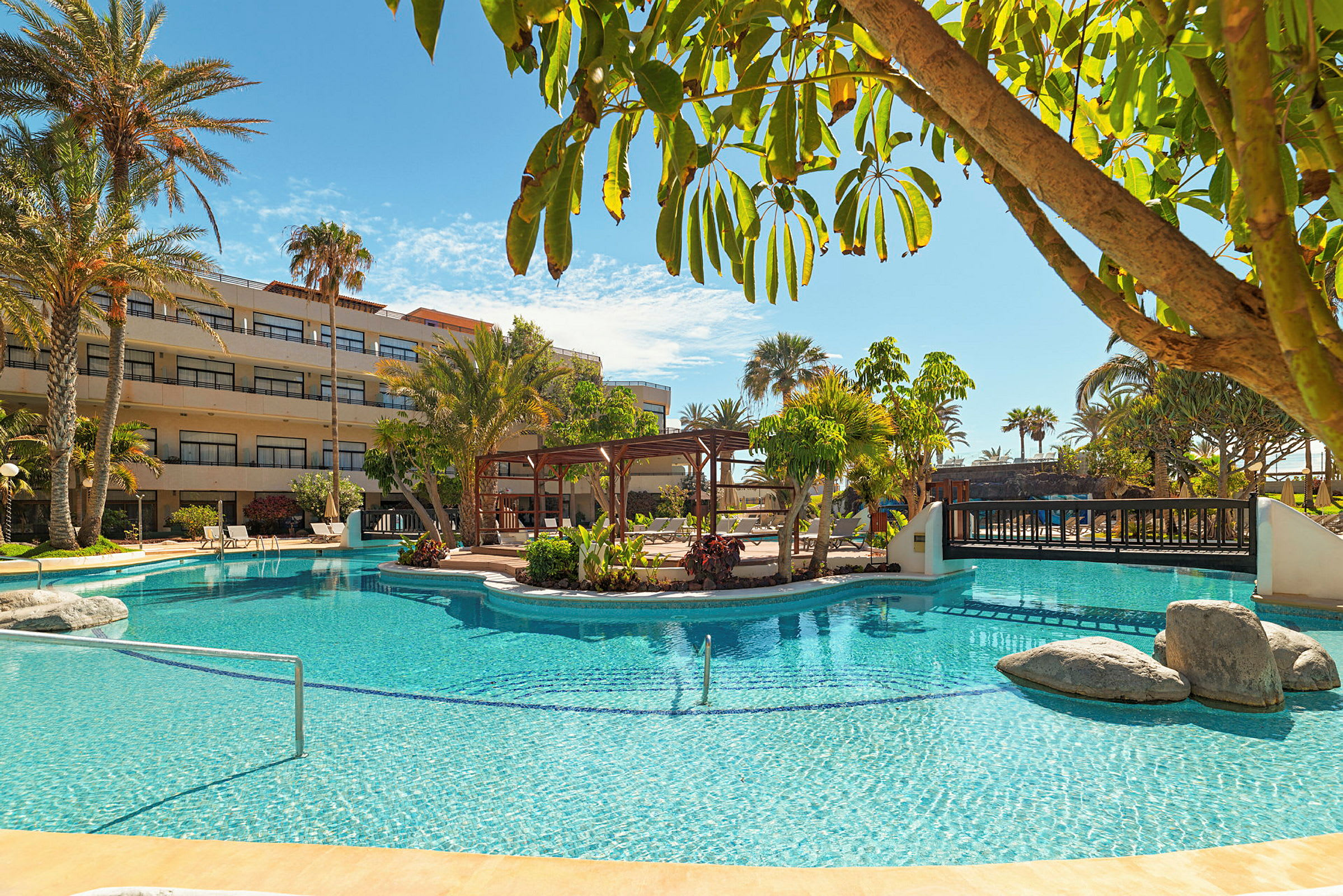 En pool ved et hotel med omgivende grøntarealer og palmer samt bygninger i baggrunden. (AI-genereret tekst)