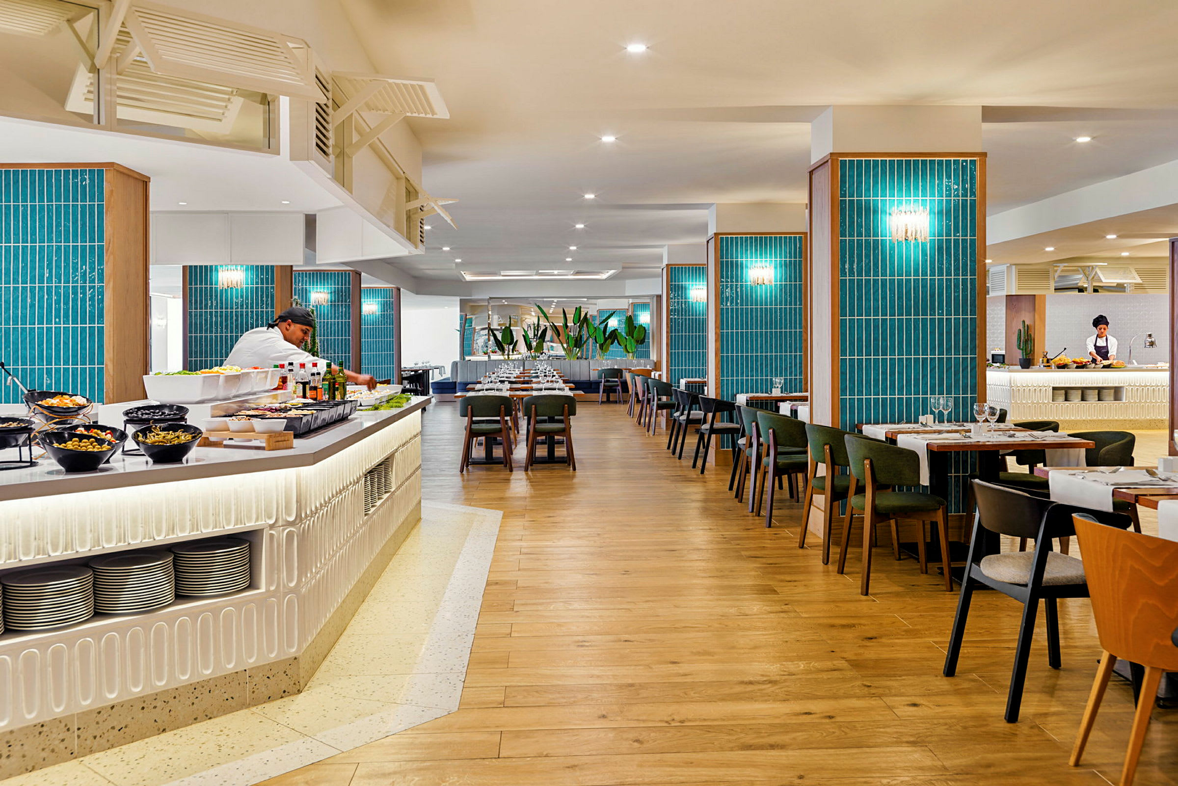 En rummelig restaurant med trægulv, blå vægdekorationer og borde med stole. En buffédisk med salater og drikkevarer ses i forgrunden, og en kok arbejder bag disken. (AI-genereret tekst)