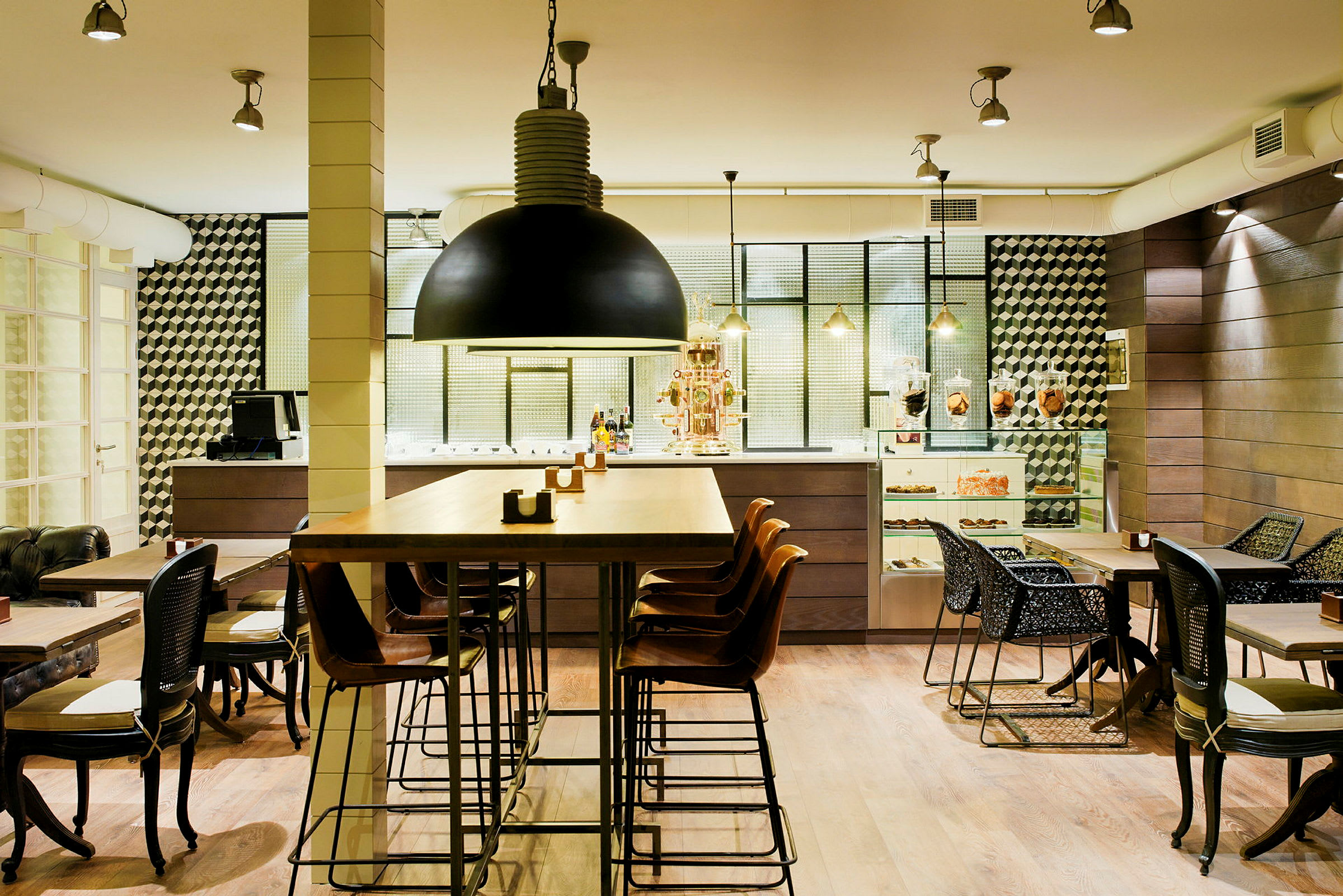 En moderne café med høje og lave borde samt stole i forskellige designs. Interiøret er stilrent med detaljer som en sort hængelampe, geometriske vægmønstre og en disk med bagværk. (AI-genereret tekst)