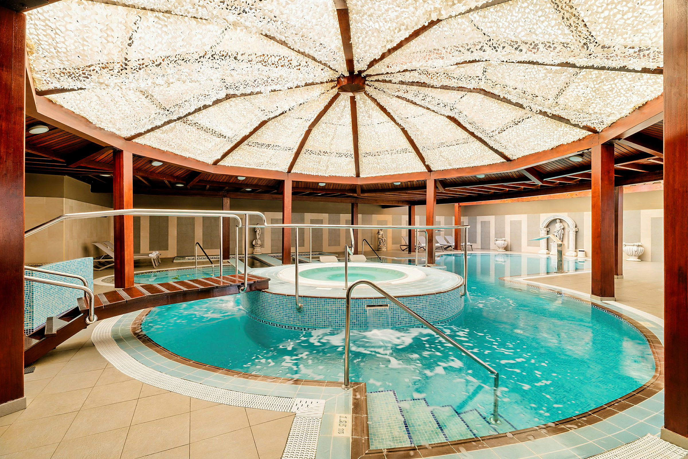 Interiør af et spa med pool og boblebad under et dekoreret loft. Træbjælker og afslapningsområder er synlige. (AI-genereret tekst)