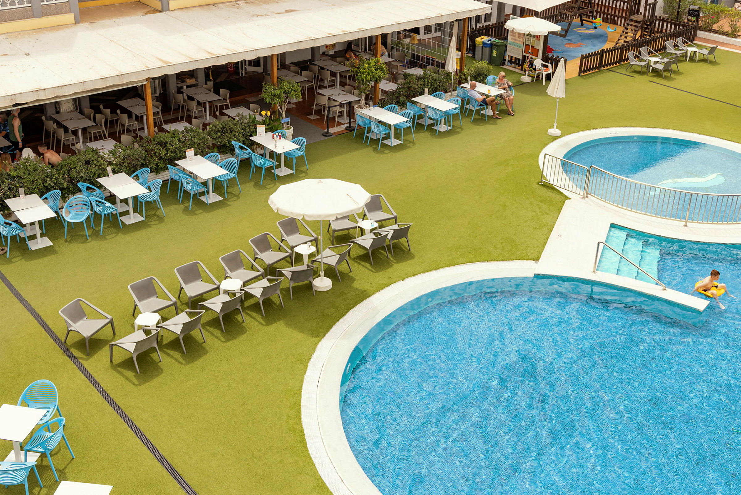 En udendørs pool med liggestole og borde rundt om samt en tilstødende restaurant. (AI-genereret tekst)