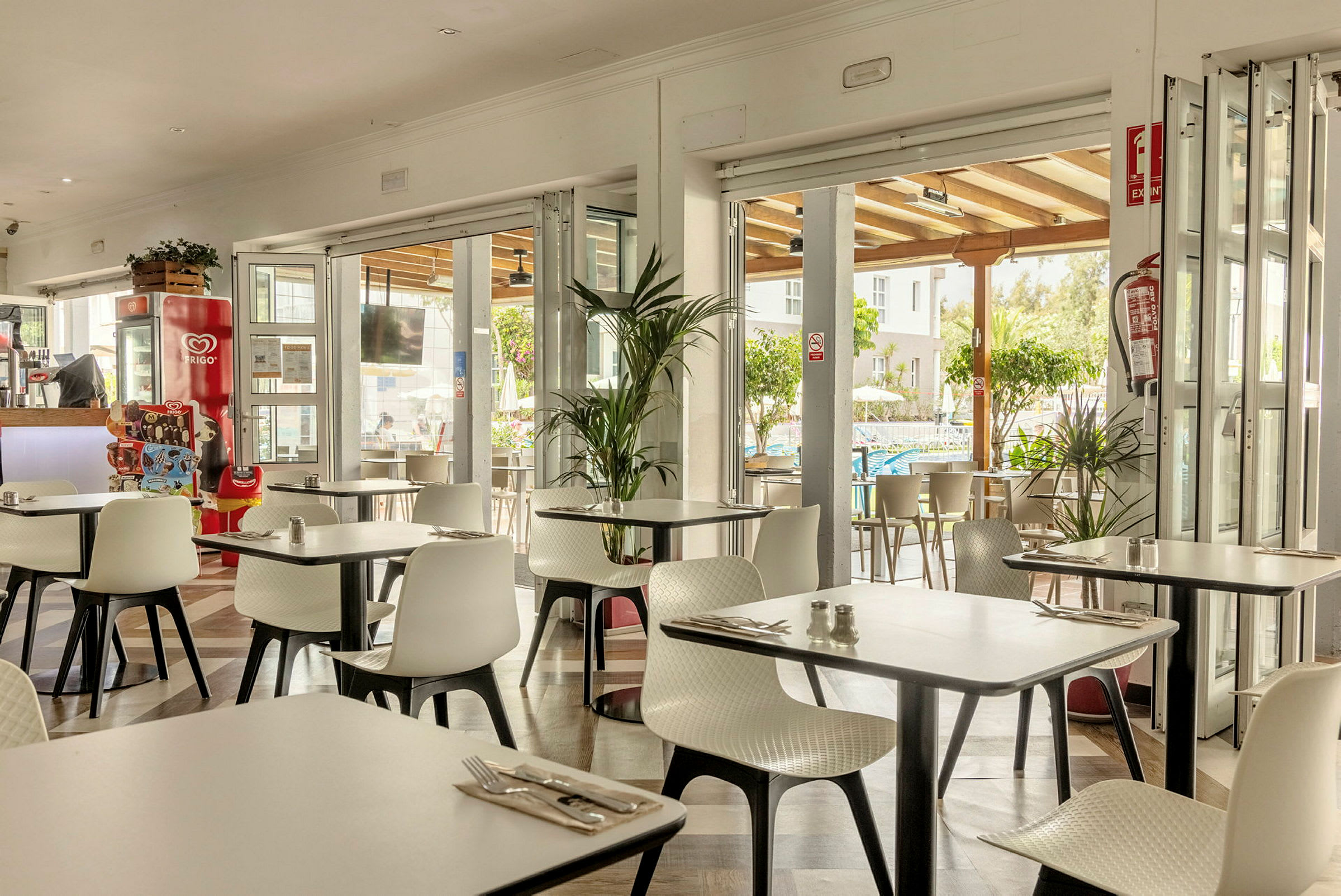 En restaurant med hvide stole og borde, åben ud mod en terrasse og omkringliggende grønne områder. (AI-genereret tekst)