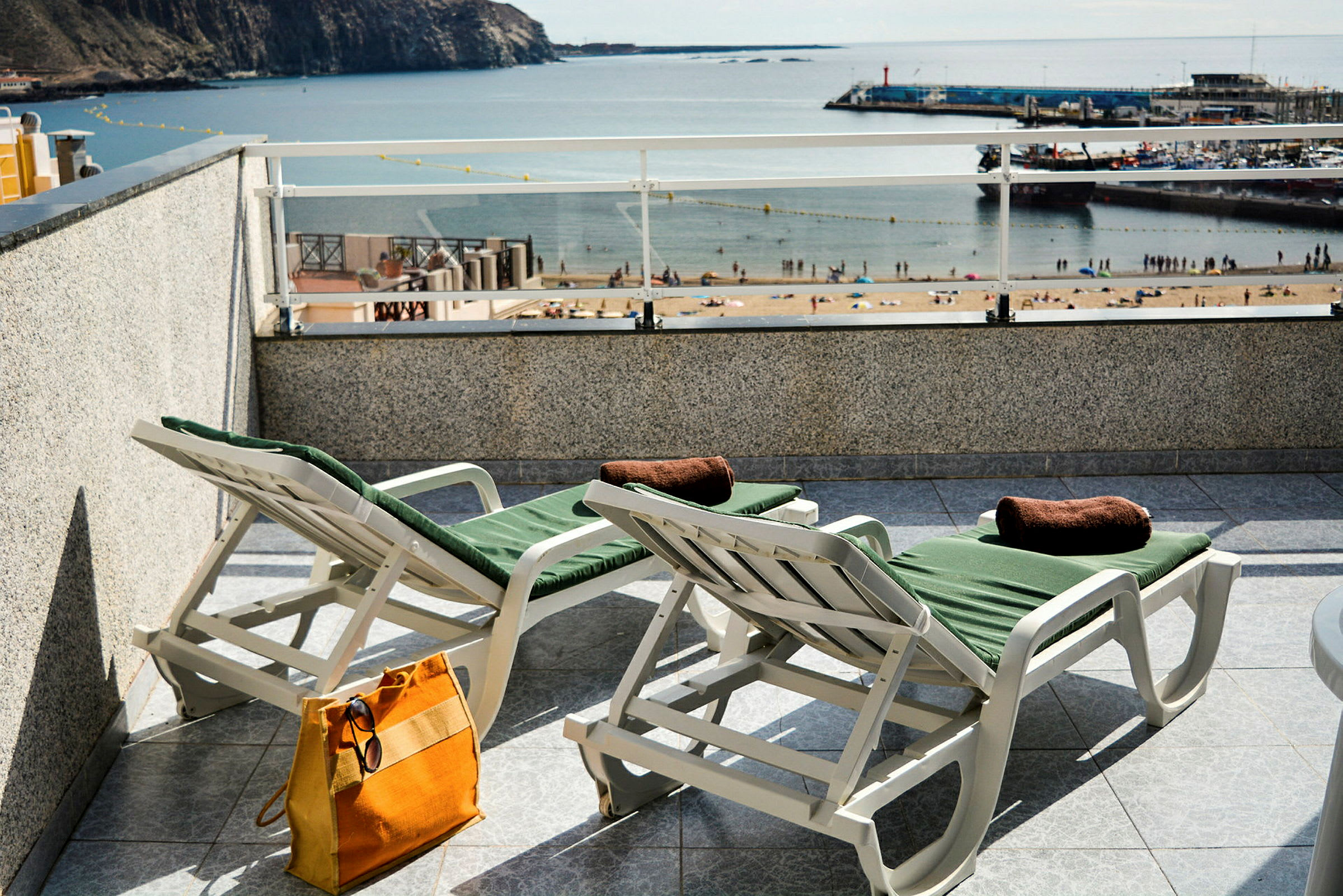 To liggestole med grønne puder på en terrasse med udsigt over en strand og havn. (AI-genereret tekst)