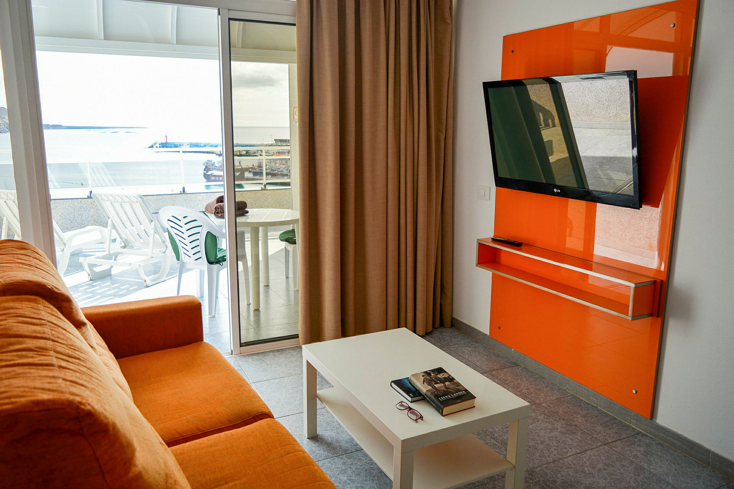 En orange sofa i et rum med et hvidt bord, et fladskærms-tv på væggen og en altan med havemøbler samt udsigt over et havneområde. (AI-genereret tekst)