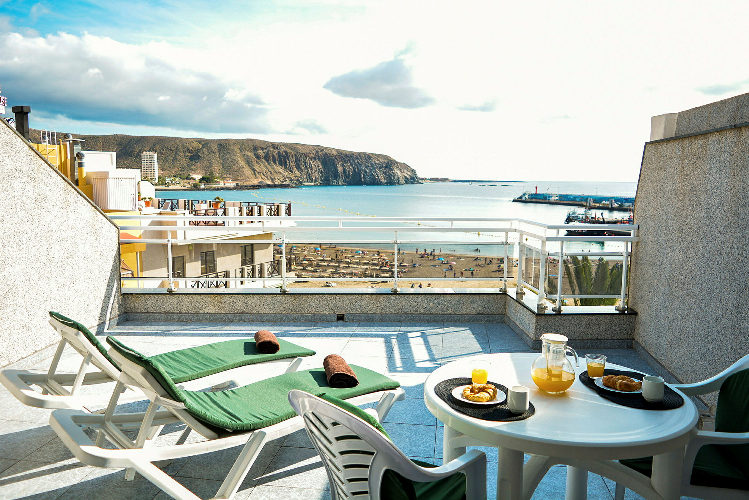 Terrasse med to solvogne og et bord med appelsinjuice og bagværk, havudsigt i baggrunden. (AI-genereret tekst)