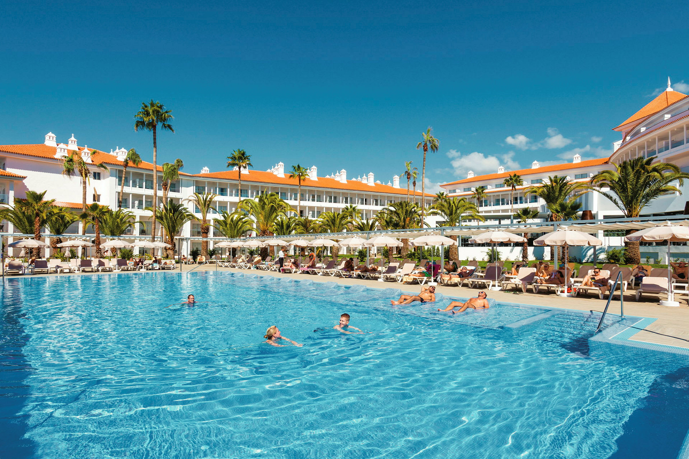 En stor udendørs pool foran et hotel med hvide bygninger og orange tage, omgivet af liggestole og palmer. (AI-genereret tekst)