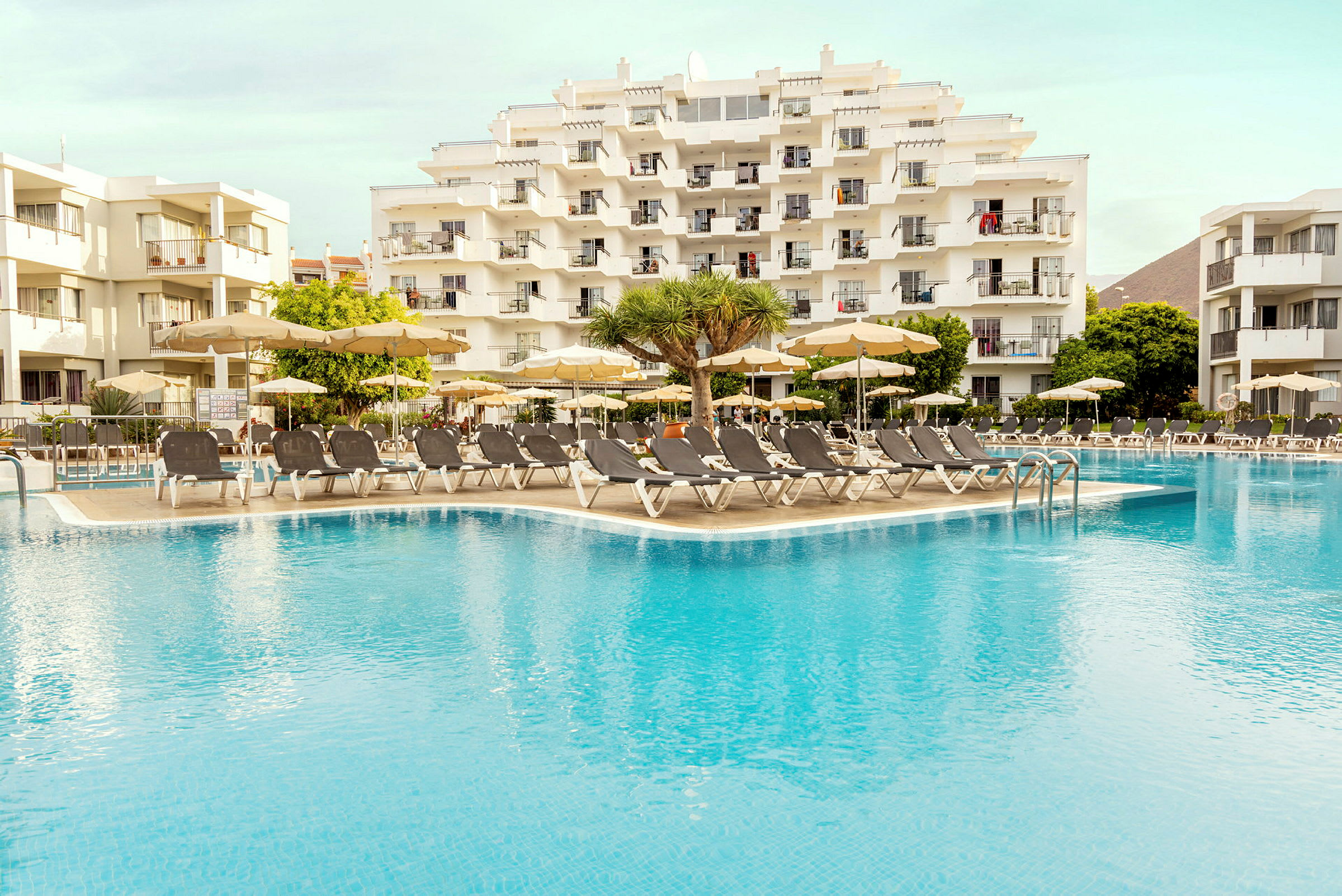 Billede af et hotelkompleks med liggestole og parasoller ved en swimmingpool. (AI-genereret tekst)