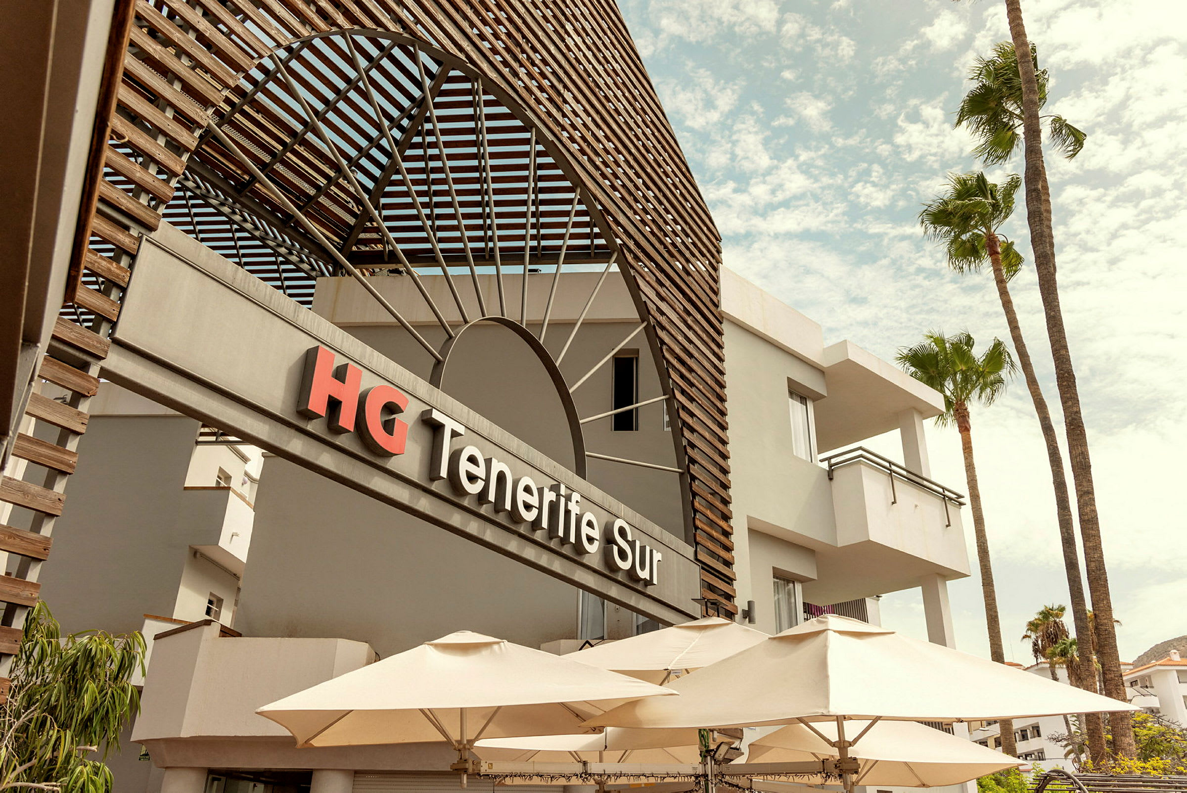 Forsiden af HG Tenerife Sur-hotellet med parasoller og palmer. (AI-genereret tekst)