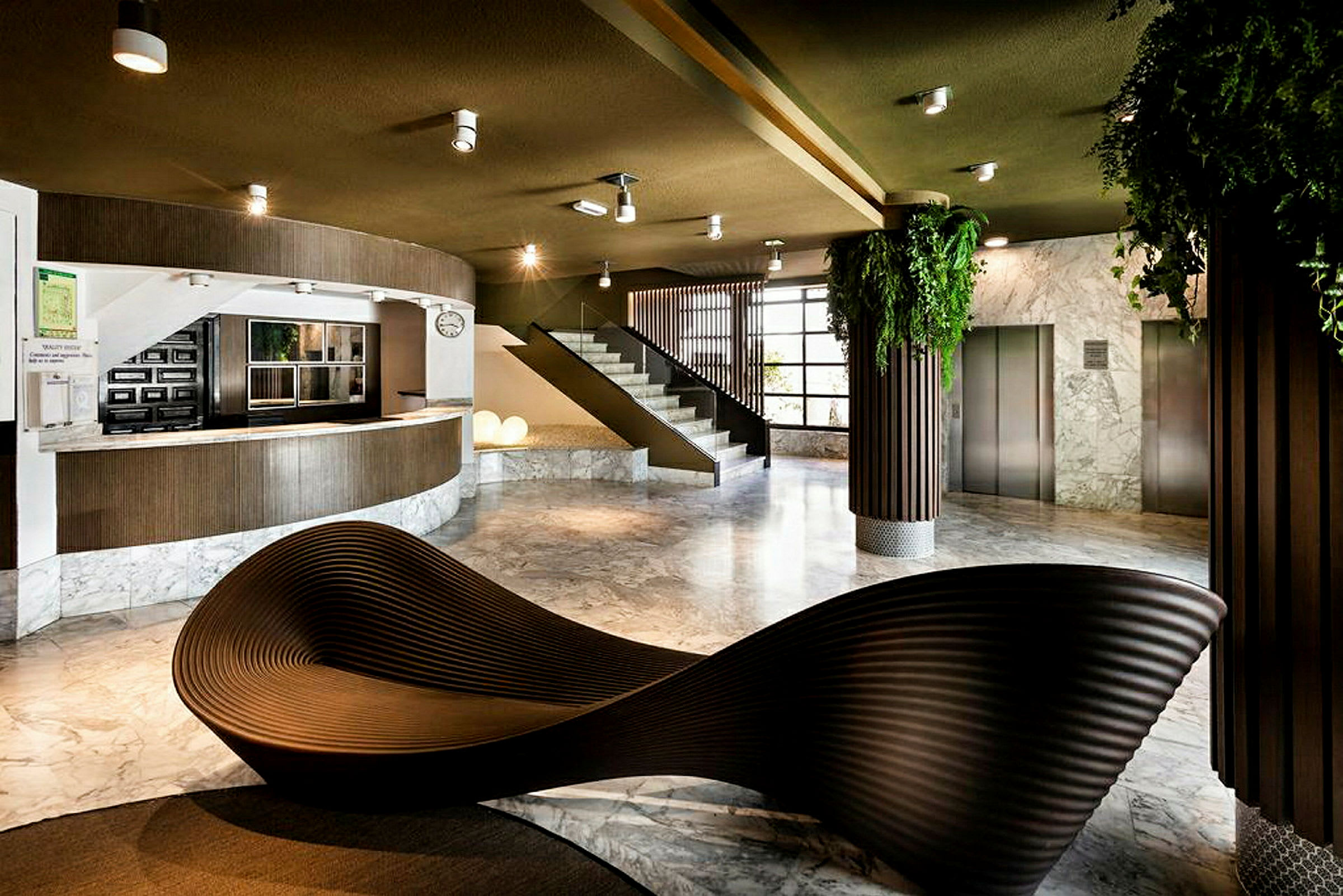 Et moderne lobbyområde med receptionsdisk, trappe og elevatorer. Indretningen inkluderer et skulpturelt siddemøbel og grønne planter. (AI-genereret tekst)