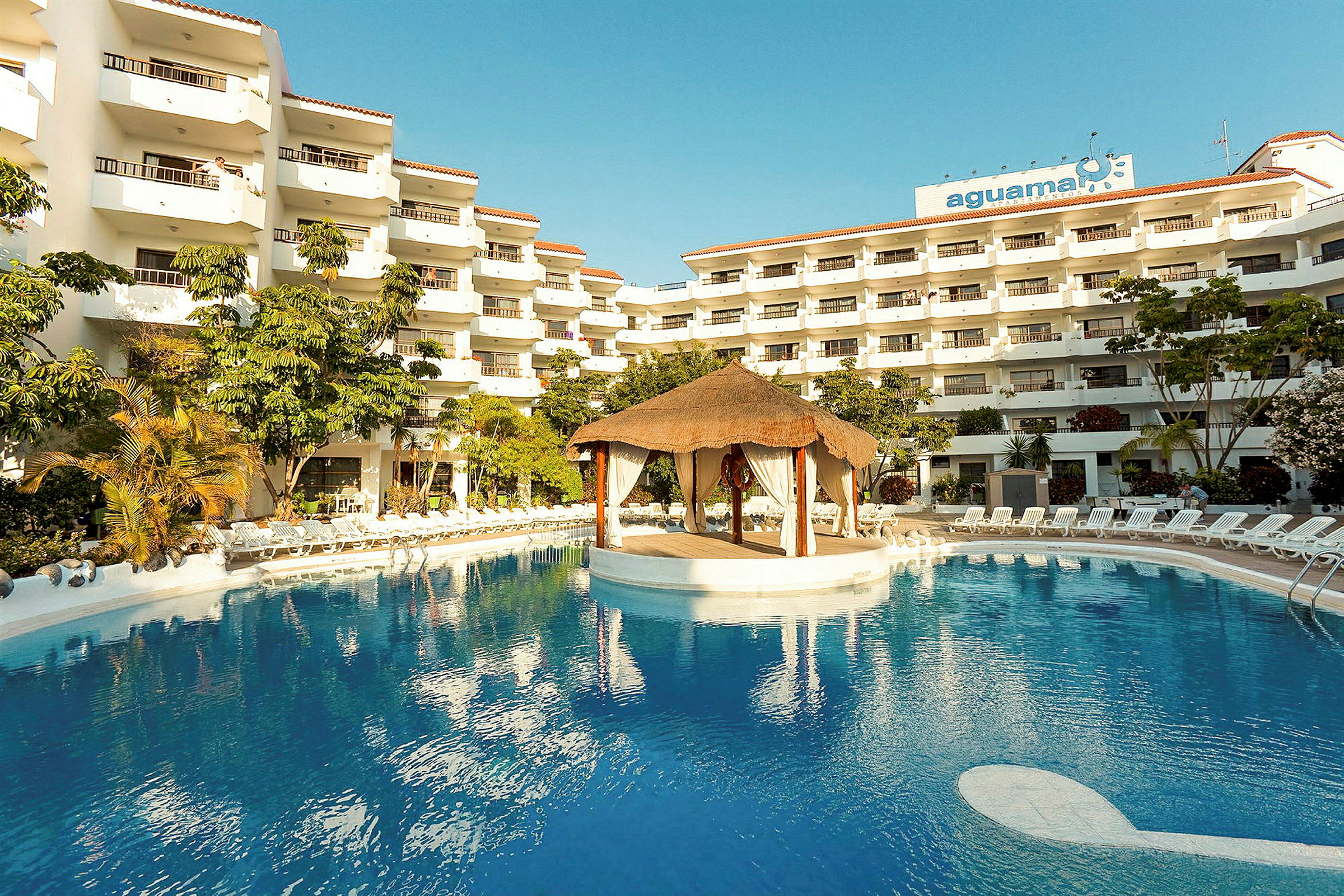 Hotelkompleks med flere etager, en stor pool og en tagehytte struktur midt i poolen. (AI-genereret tekst)