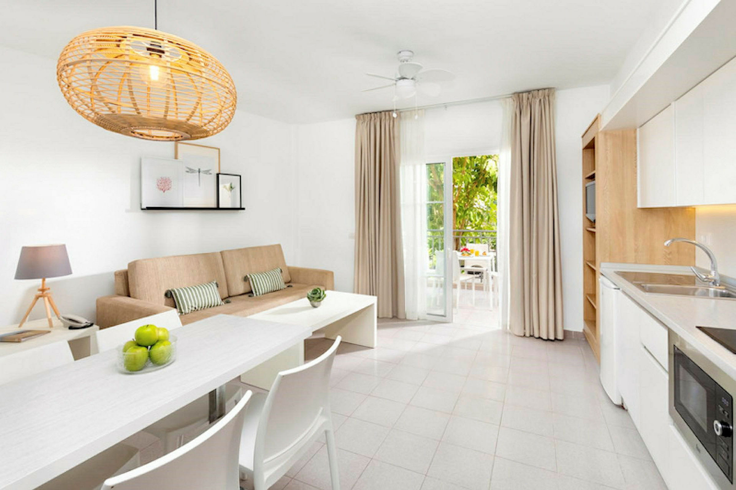 Et lyst og moderne møbleret rum med en beige sofa, hvidt spisebord og køkken samt åbne døre til en møbleret altan. (AI-genereret tekst)