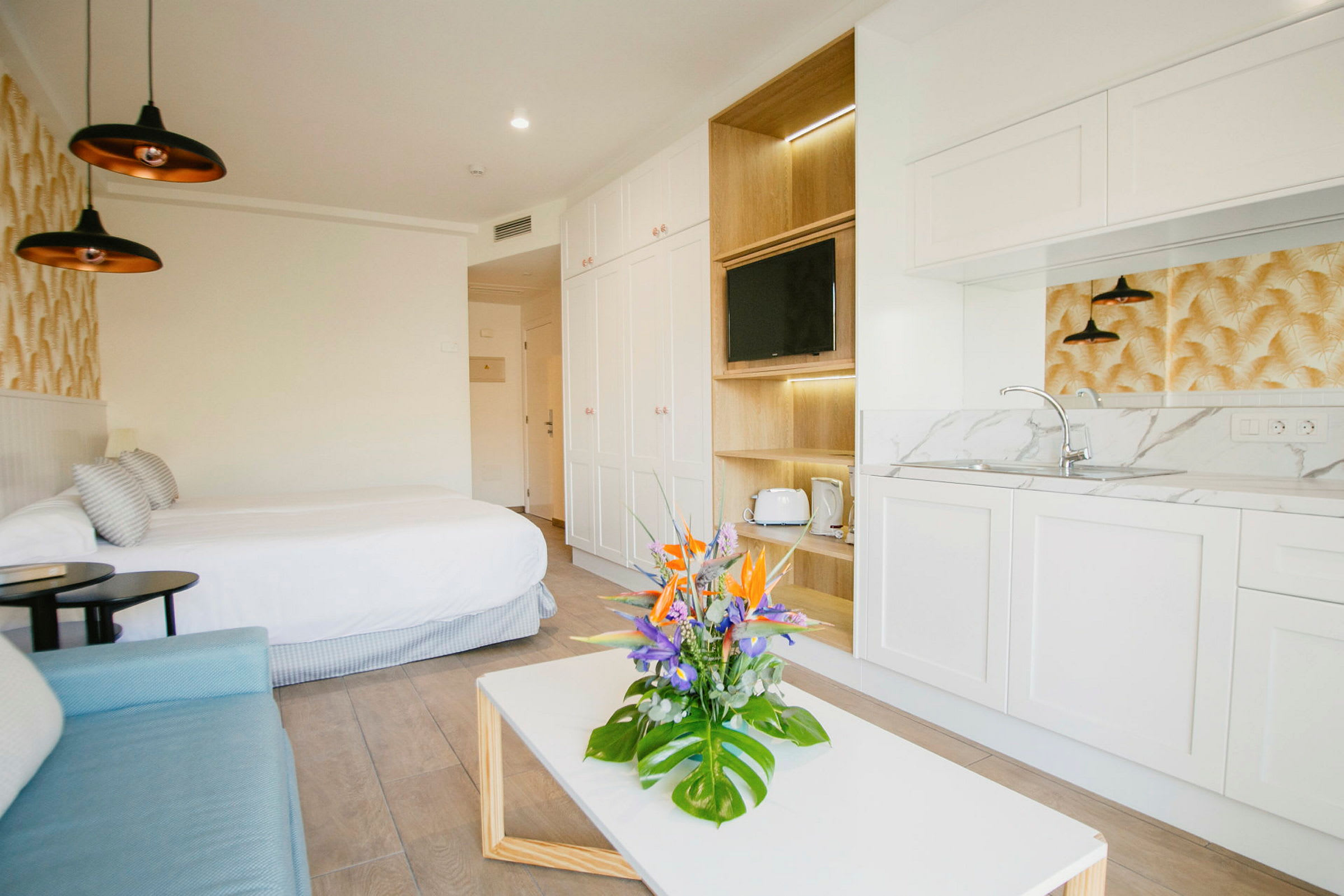 Et moderne hotelværelse med en dobbeltseng, sofa, lille køkken og et bord med en buket blomster. (AI-genereret tekst)