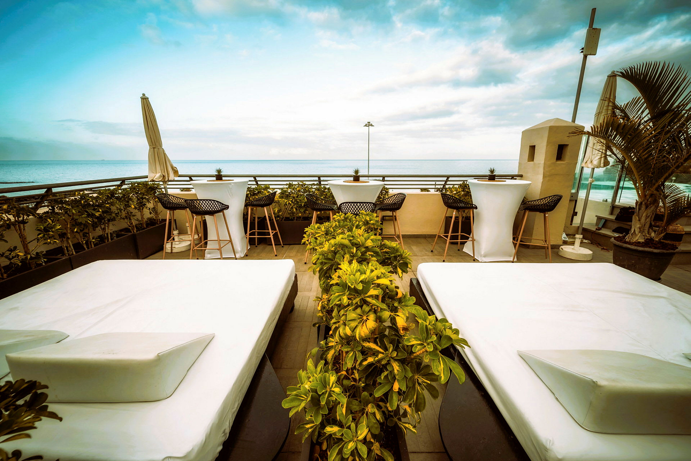En terrasse med hvide solsenge og små borde med stole. I baggrunden ses havet og blå himmel. (AI-genereret tekst)