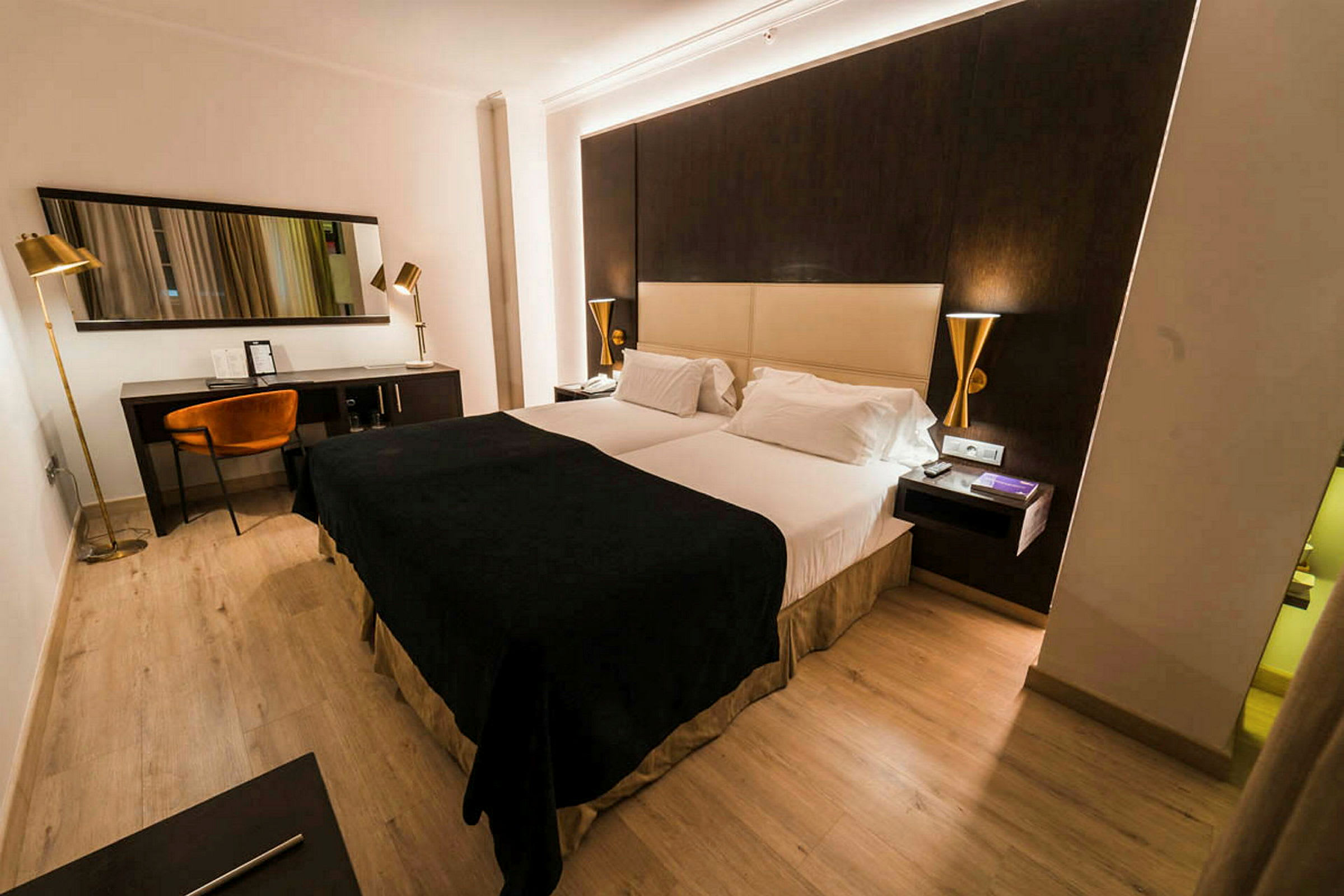 Et hotelværelse med en dobbeltseng, mørke og lyse farvetoner på møbler samt et skrivebordshjørne med stol og spejl. (AI-genereret tekst)