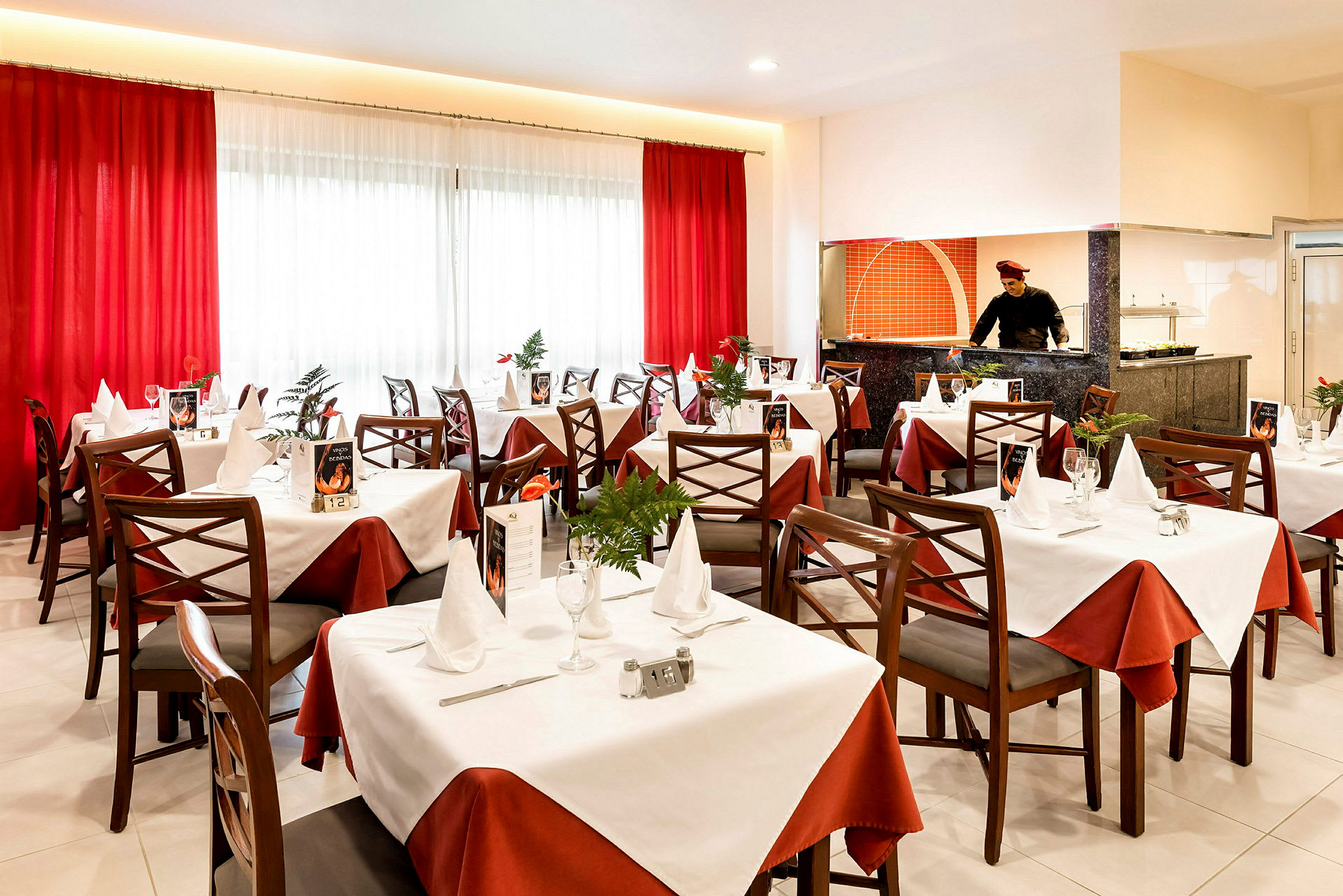 Elegant restaurant med dækkede borde, hvide og røde duge samt et åbent køkkenområde. (AI-genereret tekst)