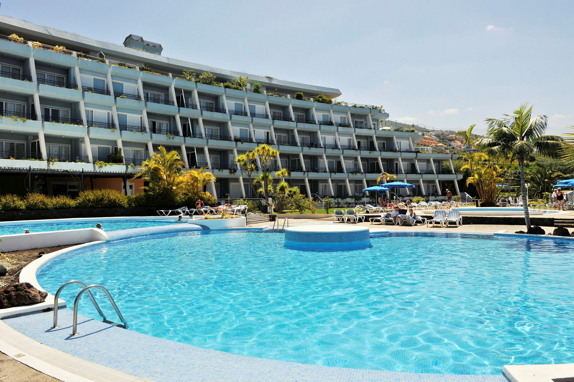 Billede af en hotelbygning med flere etager, swimmingpool og liggestole i forgrunden. (AI-genereret tekst)
