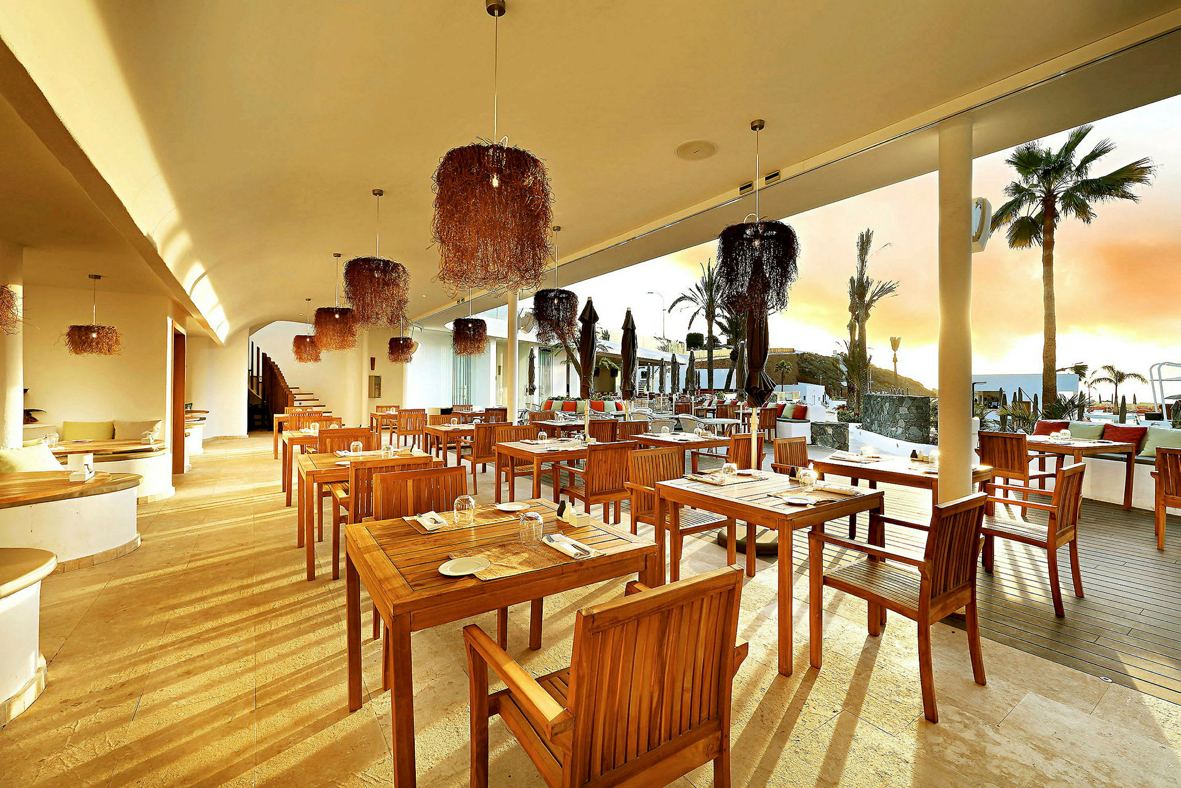 En udendørs restaurant med træborde og stole, moderne indretning med hængende lamper, solnedgang i baggrunden. (AI-genereret tekst)