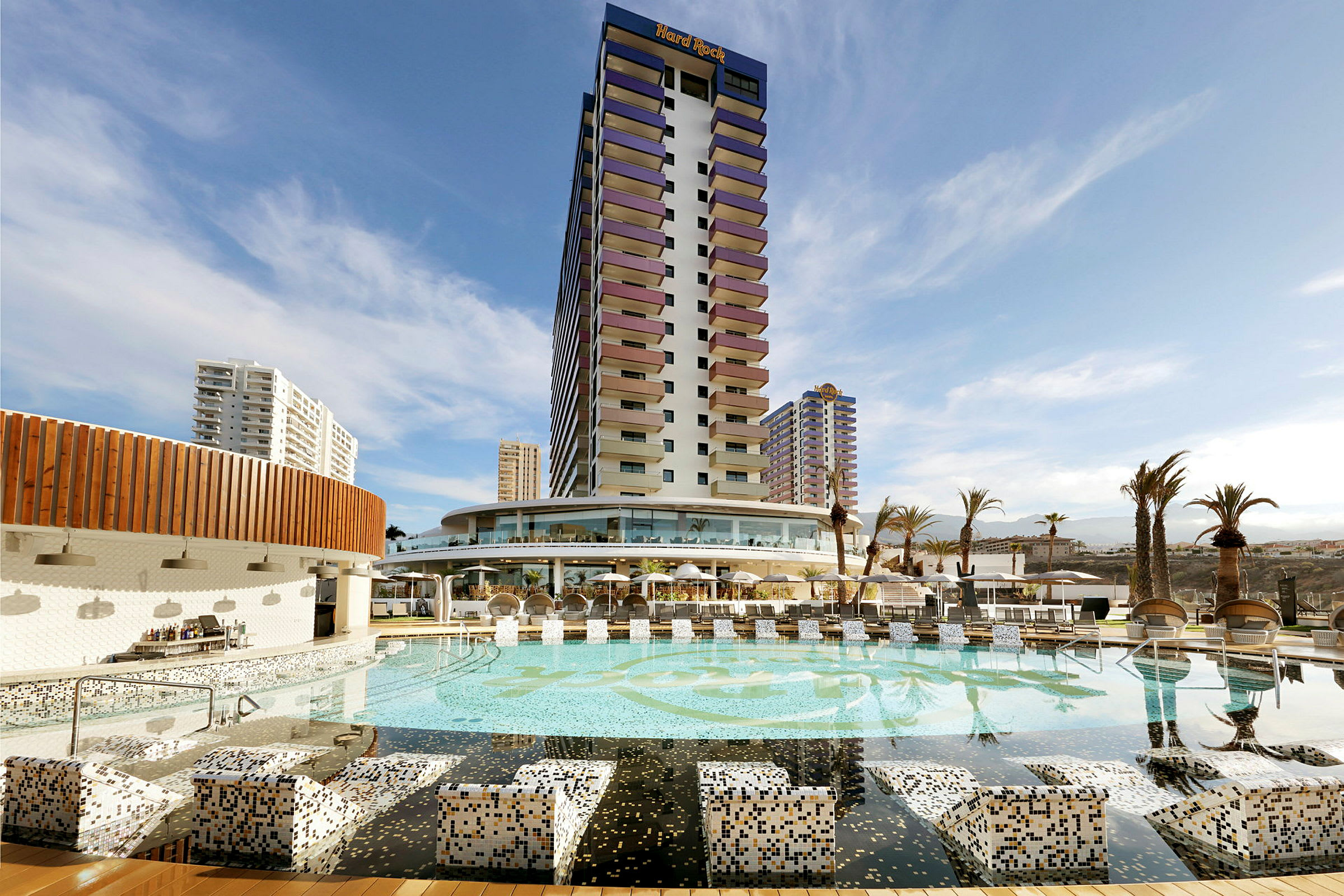 Et hotel med en stor pool og omkringliggende bygninger under en blå himmel. (AI-genereret tekst)