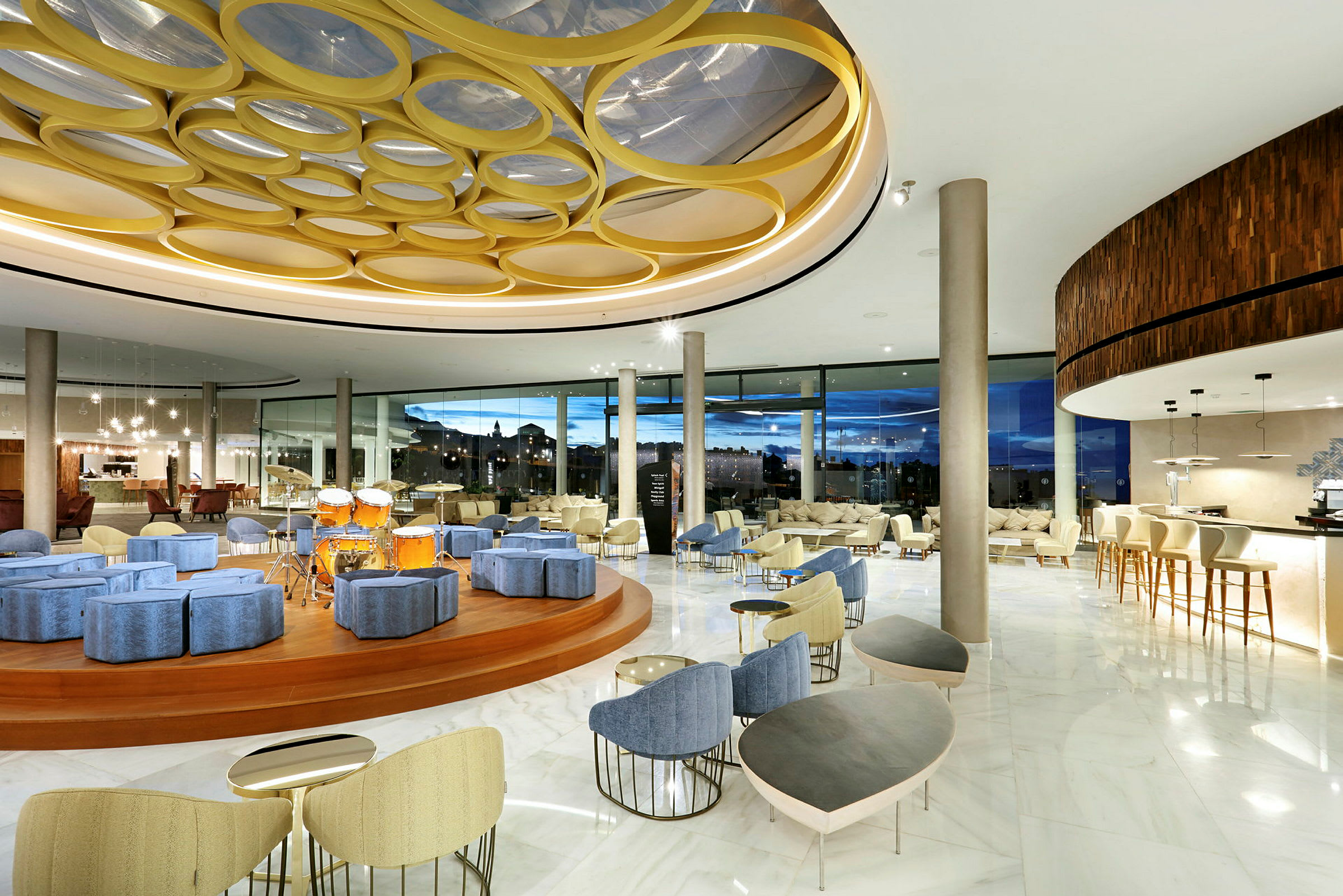 Interiør af en moderne hotellobby med siddegrupper, en scene med trommer og store panoramavinduer. (AI-genereret tekst)