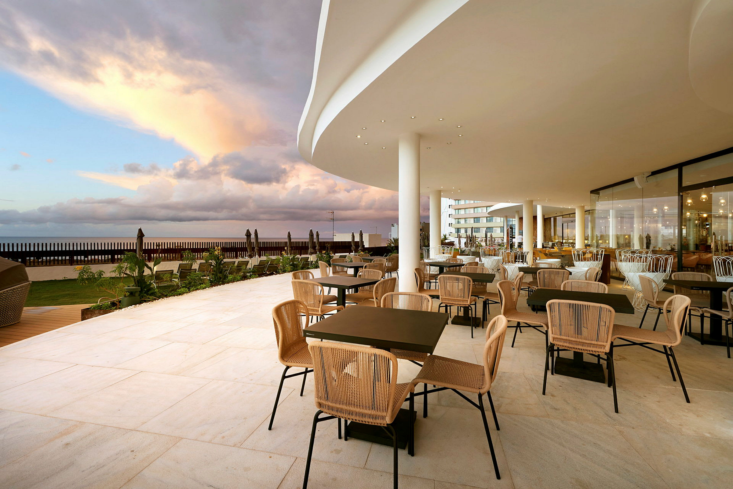 En restaurantterrasse med borde og stole samt udsigt over havet under en skyet himmel. (AI-genereret tekst)