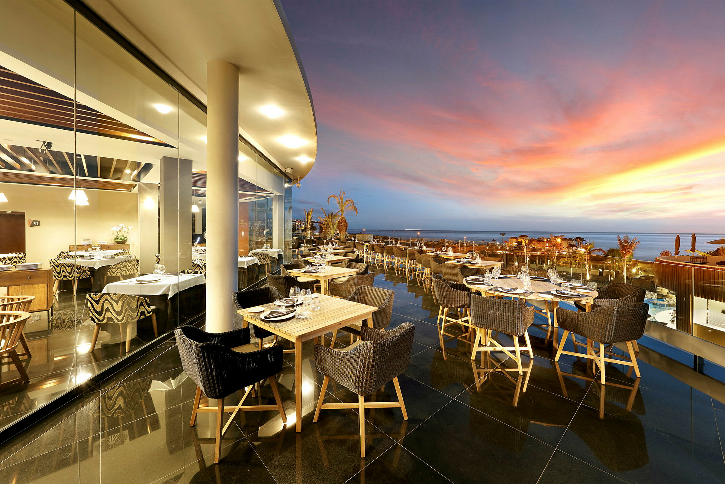 En udendørs restaurant med borde og stole ved en glasvæg med udsigt over en naturlig solnedgang. (AI-genereret tekst)