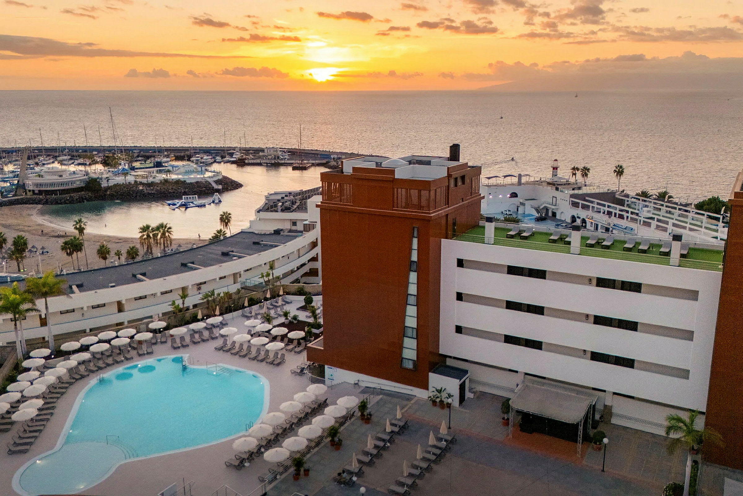 Billede af et hotel med poolområde i forgrunden og en solnedgang over en marina i baggrunden. (AI-genereret tekst)