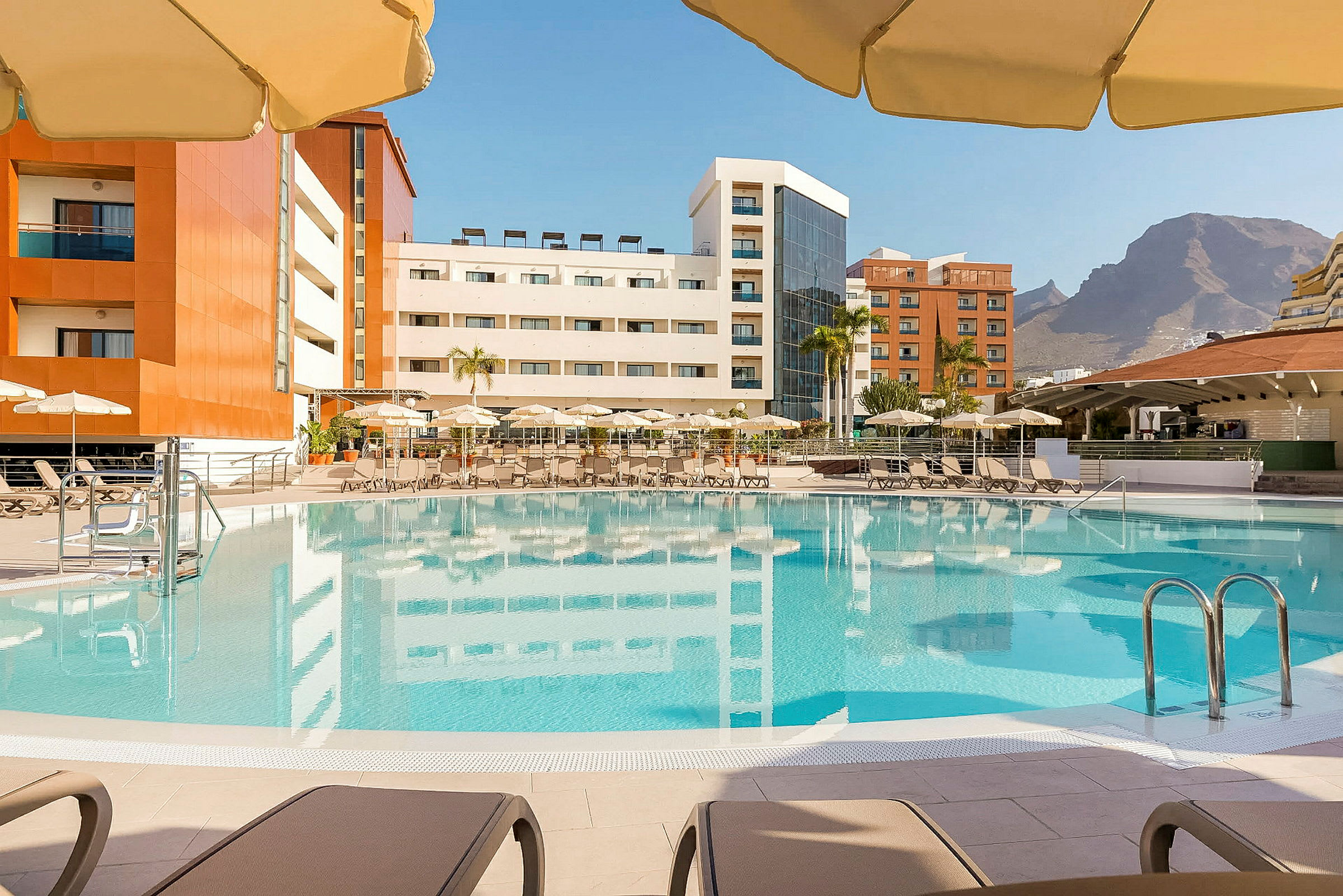 En pool omgivet af liggestole og parasoller, med hotelbygninger i baggrunden og bjerge i det fjerne. (AI-genereret tekst)