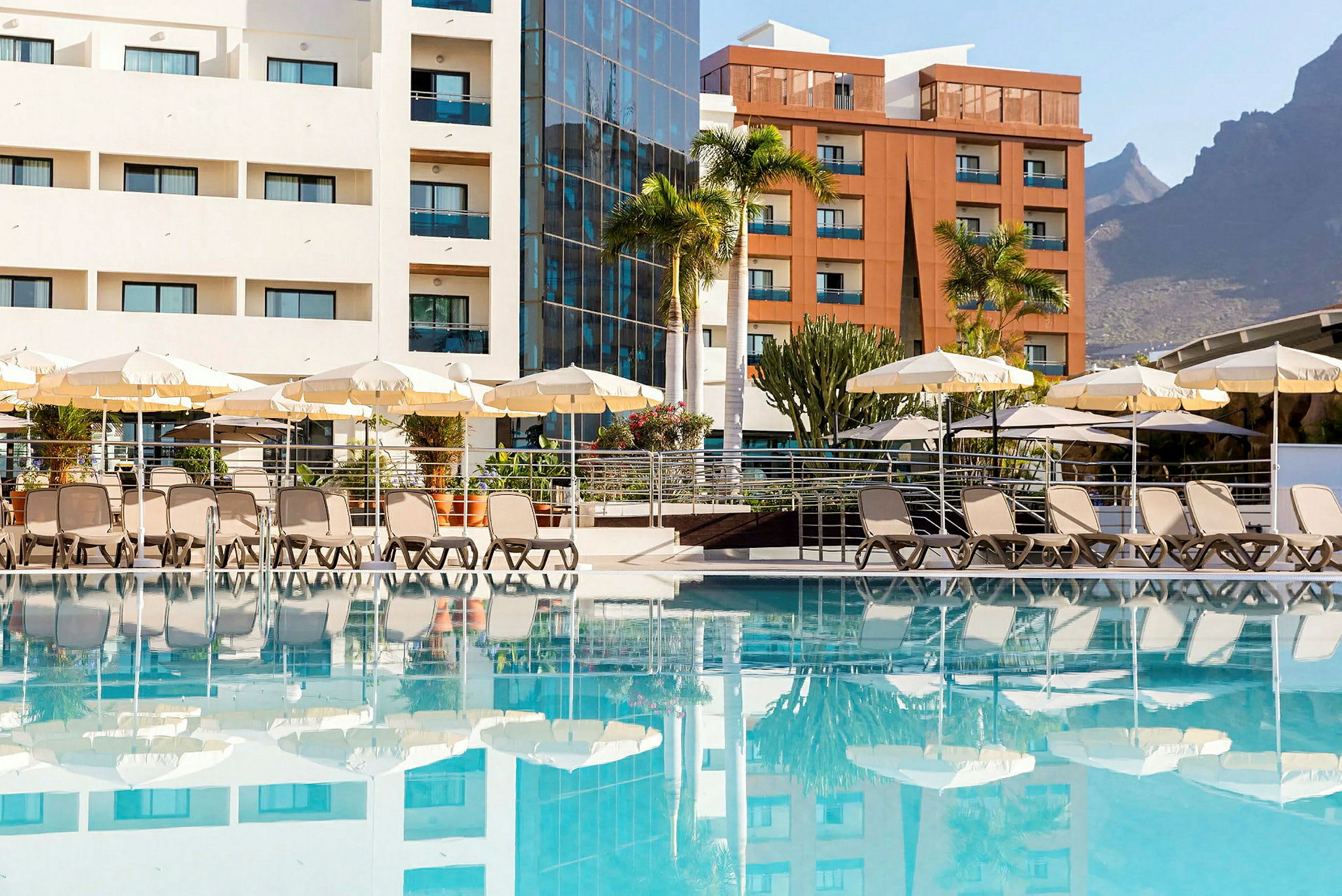 Udendørs swimmingpool med liggestole og parasoller foran hotelbygninger. (AI-genereret tekst)