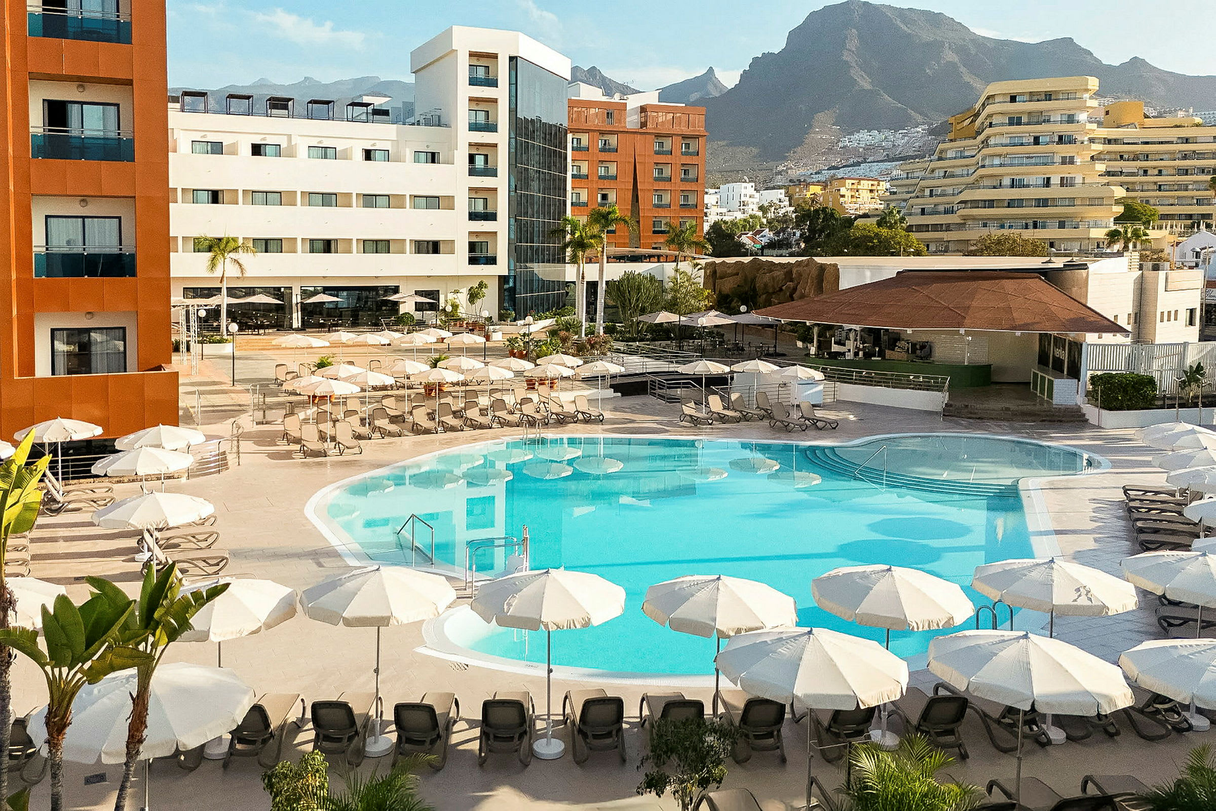 Hotel med poolområde omgivet af parasoller og liggestole samt omkringliggende bygninger og bjerge i baggrunden. (AI-genereret tekst)