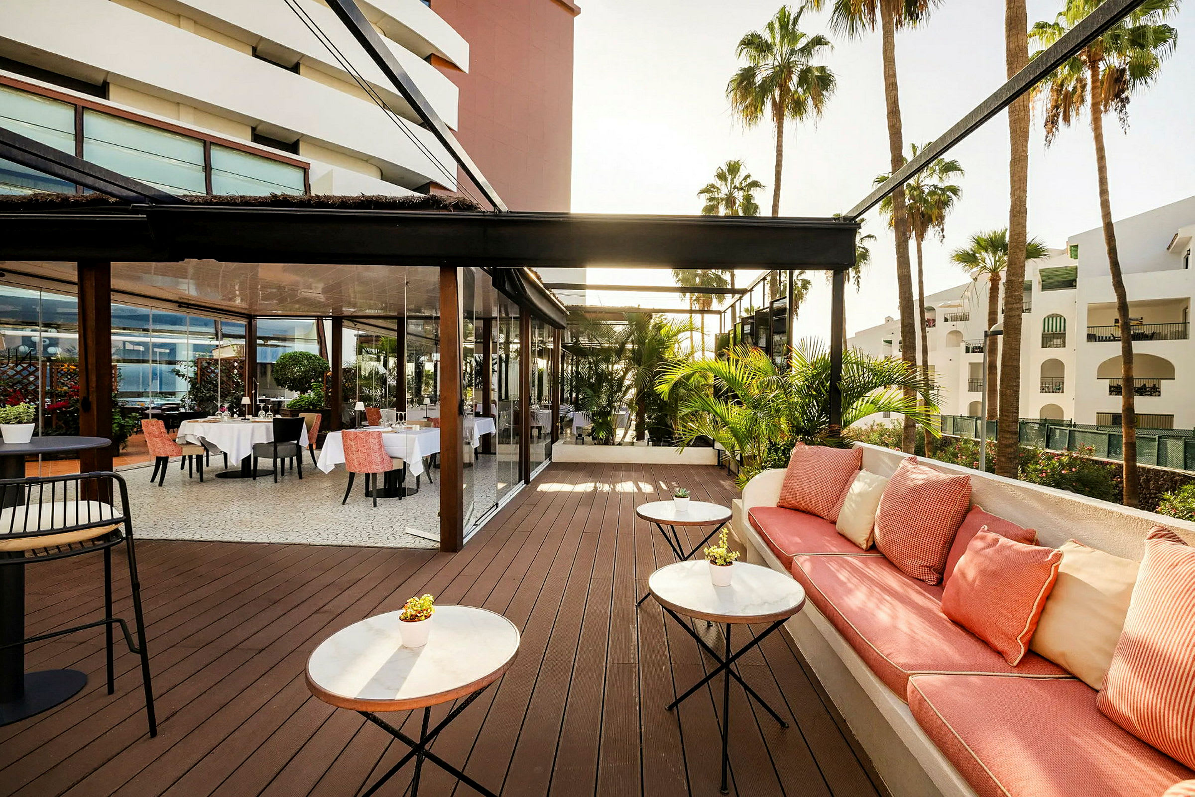En udendørs terrasse med borde og stole samt en siddeplads med puder. Palmer og hvide bygninger ses i baggrunden. (AI-genereret tekst)