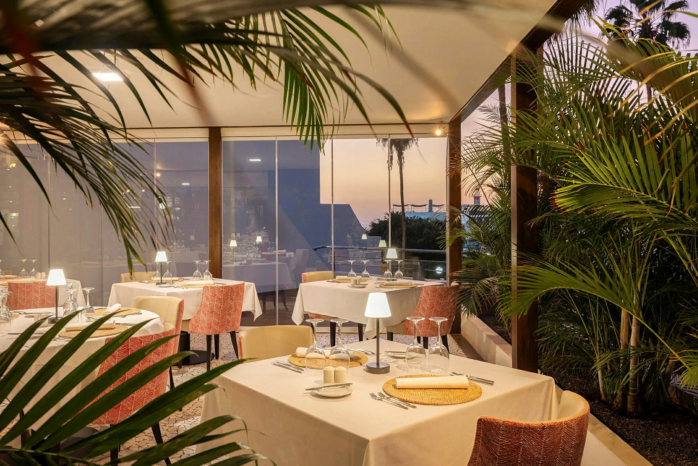 En elegant restaurant med dækkede borde indendørs omgivet af grønt, med solnedgang i baggrunden. (AI-genereret tekst)