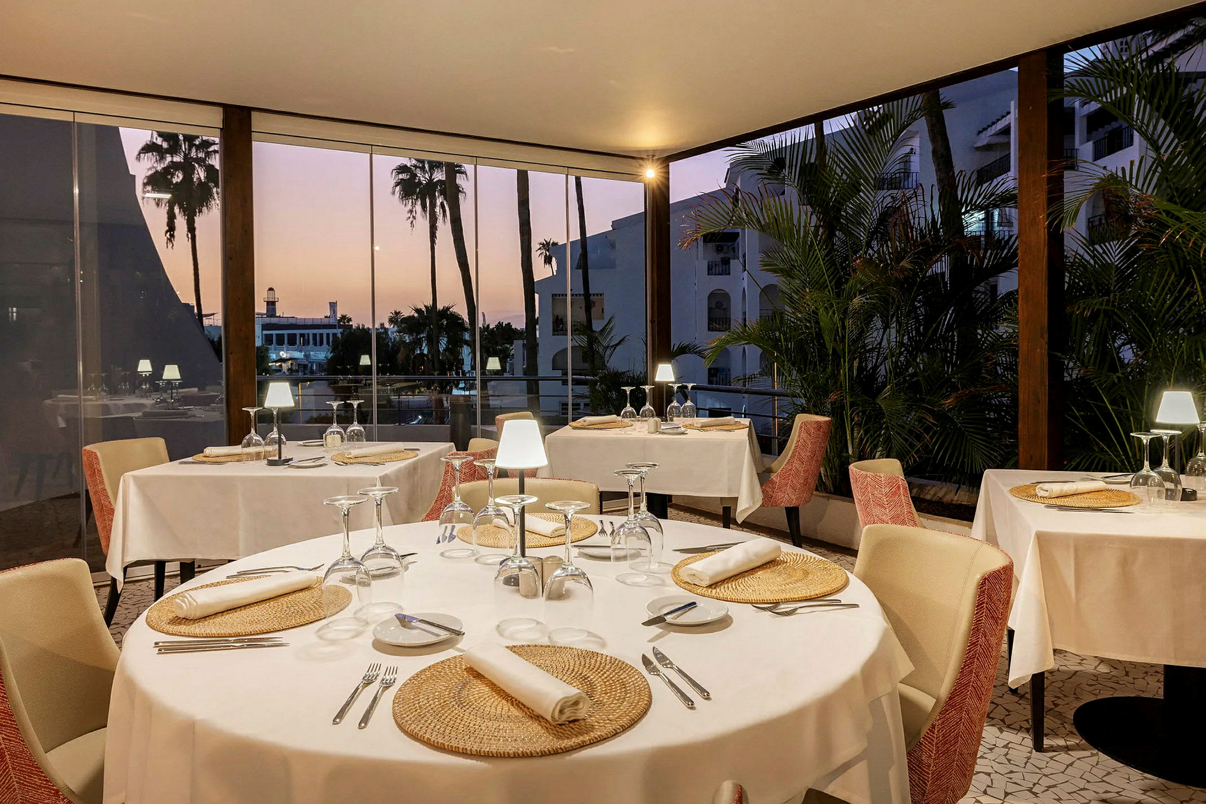 Inde i en restaurant med dækkede borde og udsigt til palmer gennem store vinduer. (AI-genereret tekst)