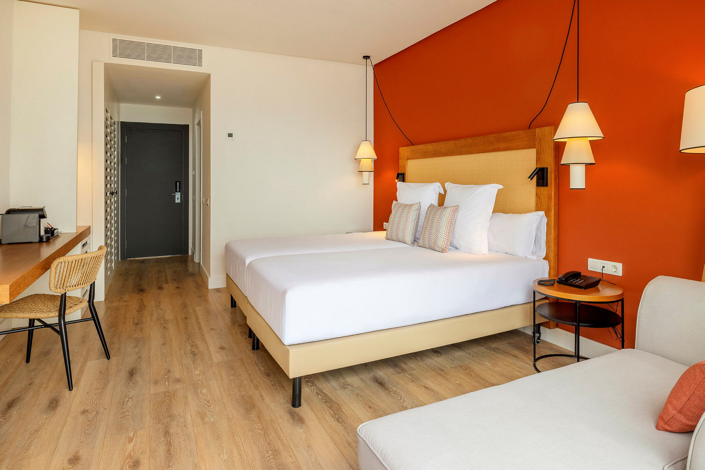 Et hotelværelse med en orange væg, en dobbeltseng, skrivebord og stole. (AI-genereret tekst)