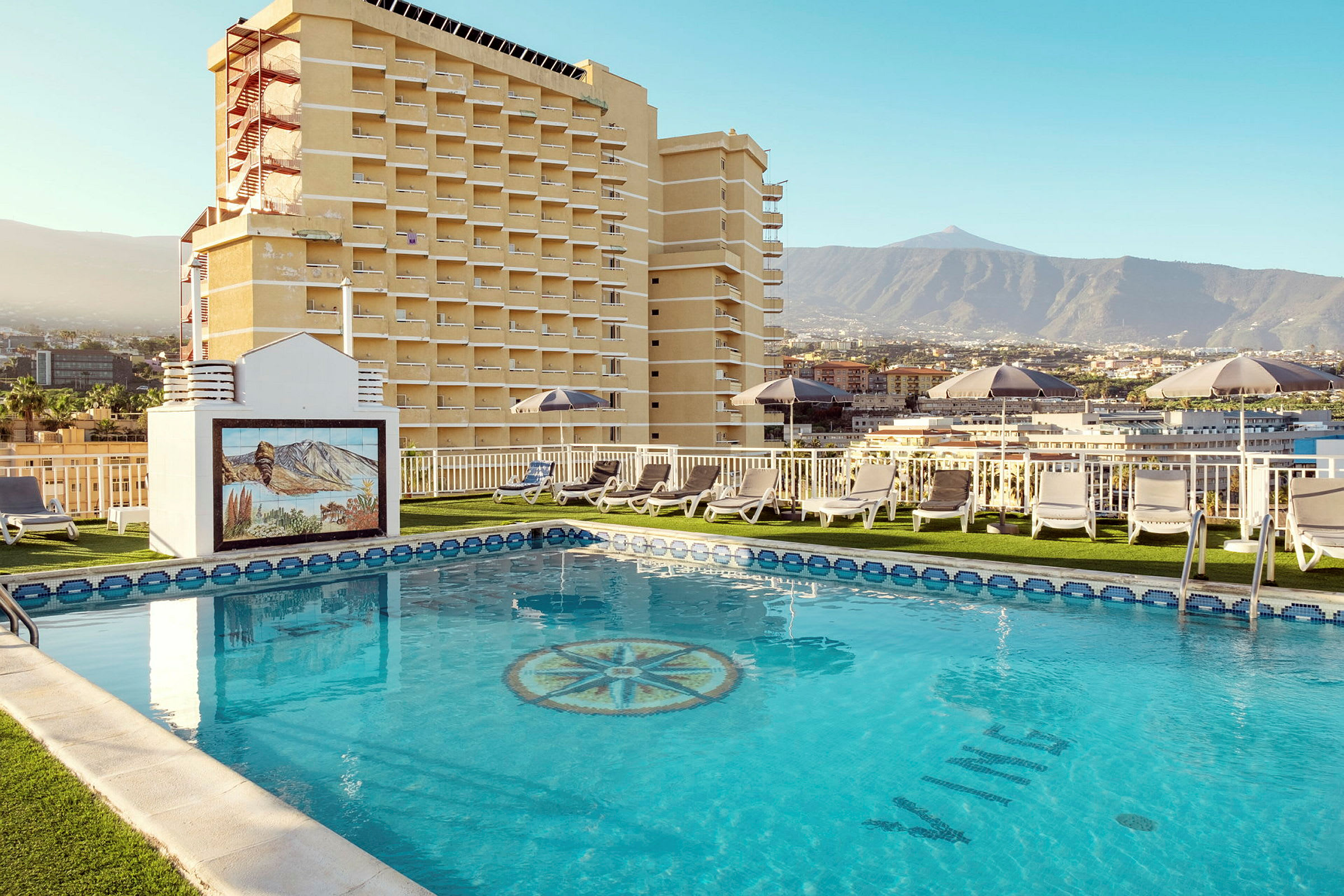 En hotelpool med solvogne og udsigt til bjerge og bygninger. (AI-genereret tekst)