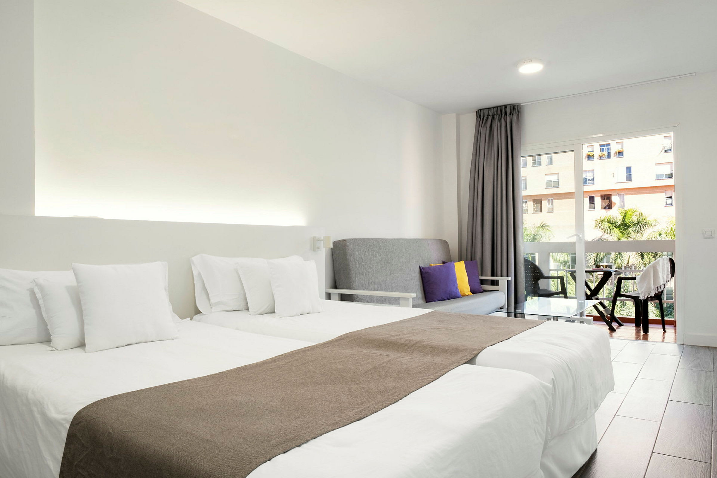 Et hotelværelse med to enkeltsenge med hvidt sengetøj, en grå sofa med farvede puder og en balkon med udsigt. (AI-genereret tekst)