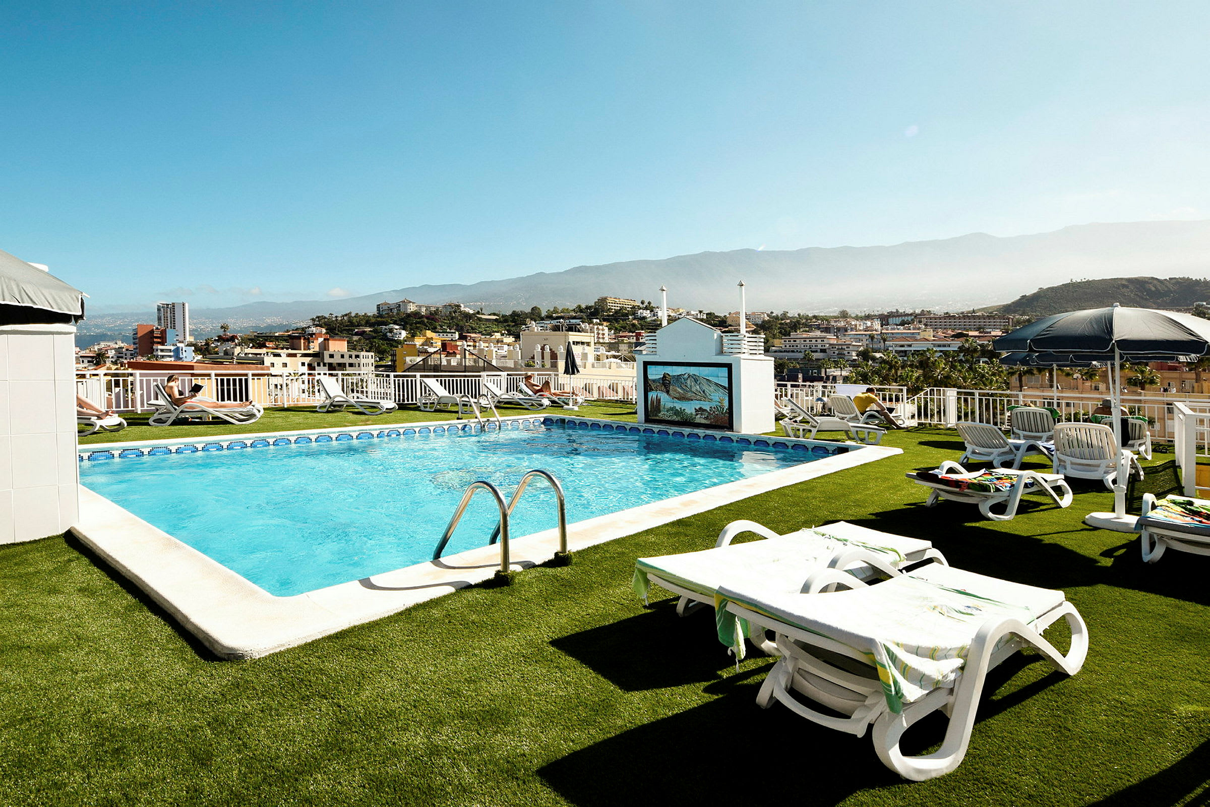 Billede af en udendørs swimmingpool med liggestole omkring, beliggende på en terrasse med udsigt over en by. (AI-genereret tekst)