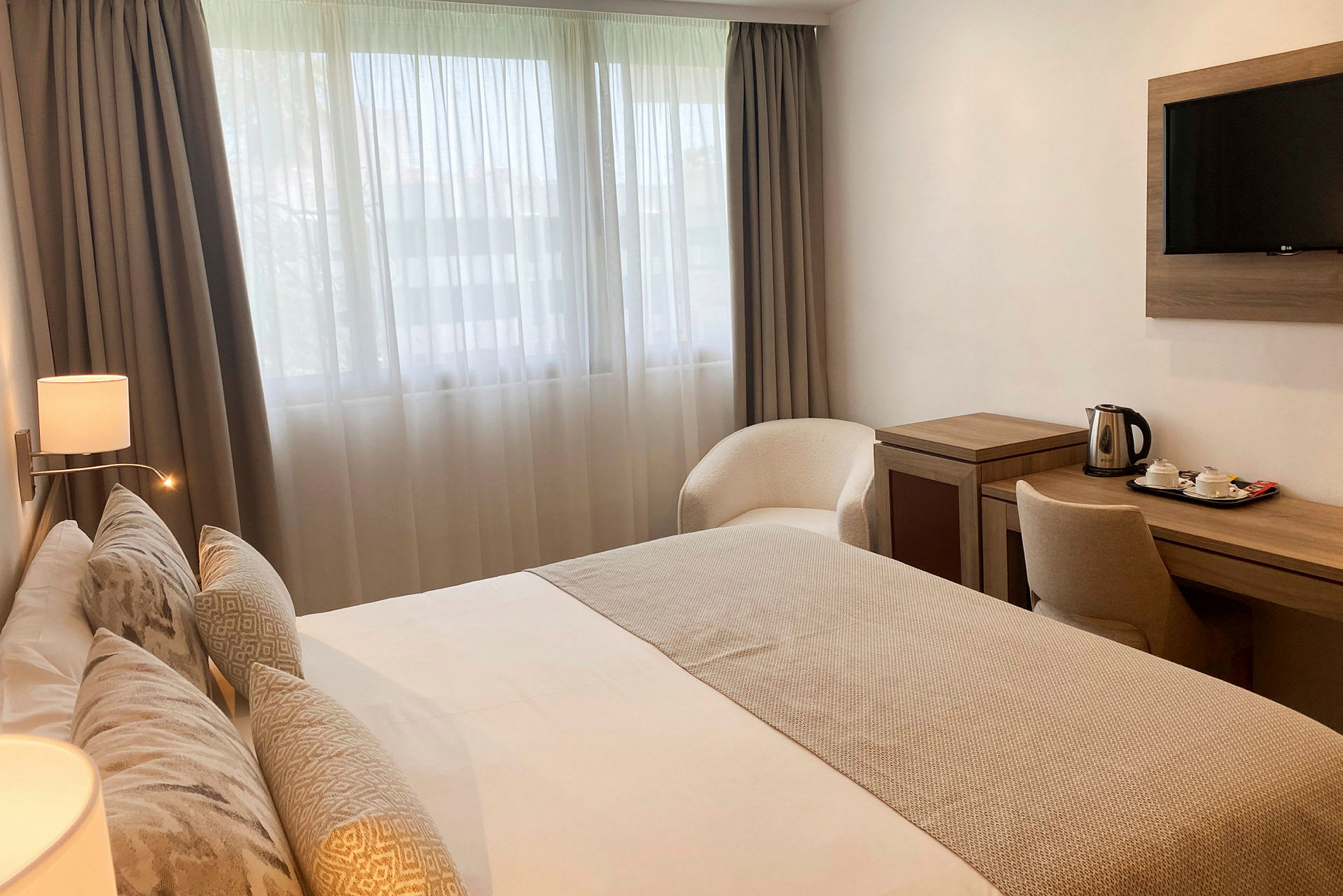 Hotelværelse med en dobbeltseng, skrivebord med en elkedel og tekopper, samt en lænestol placeret nær vinduet. (AI-genereret tekst)