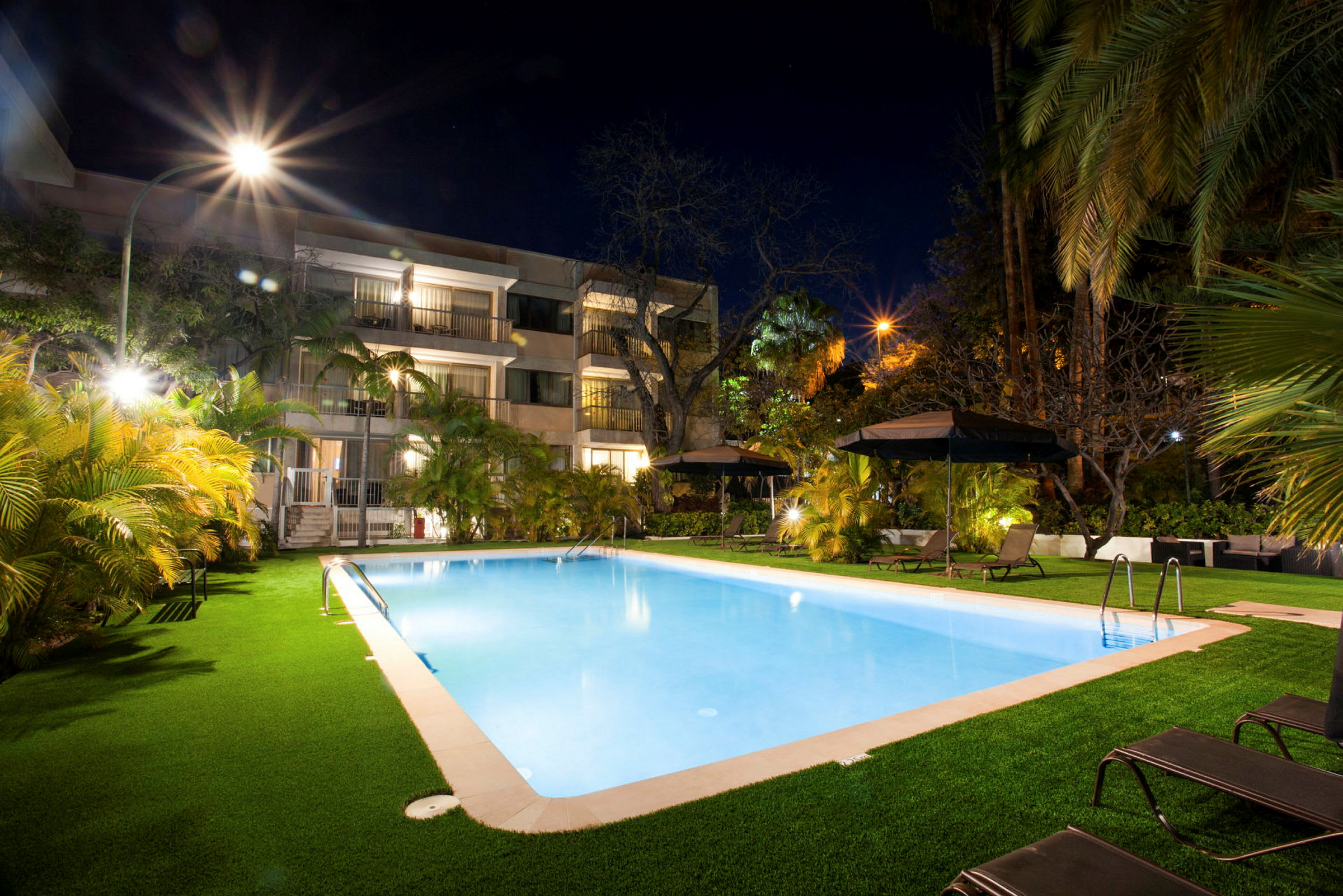 En oplyst swimmingpool om aftenen ved et hotel med omkringliggende vegetation og liggestole. (AI-genereret tekst)