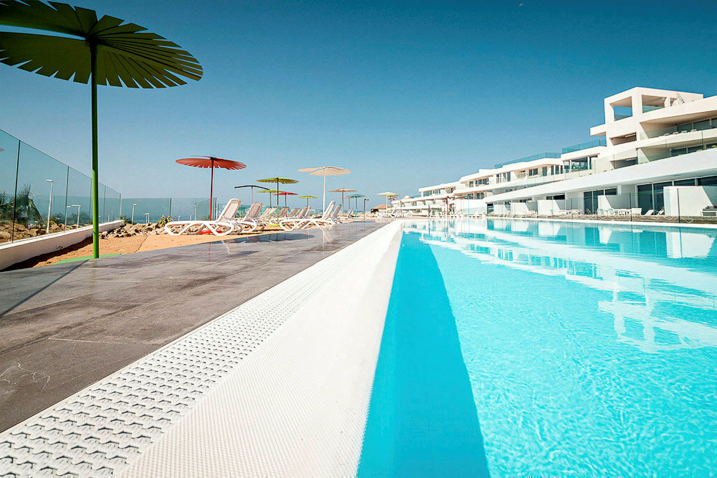 En stor swimmingpool med omkringliggende liggestole og parasoller udenfor. (AI-genereret tekst)