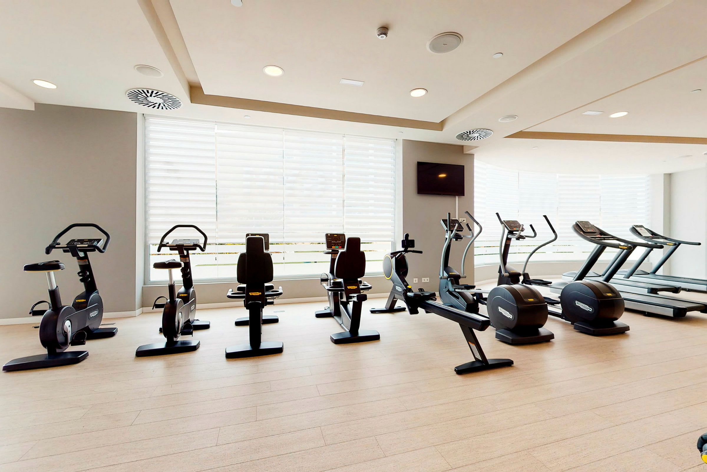 Et træningsrum med fitnessmaskiner såsom stationære cykler, løbebånd og elliptiske maskiner. Store vinduer i baggrunden. (AI-genereret tekst)