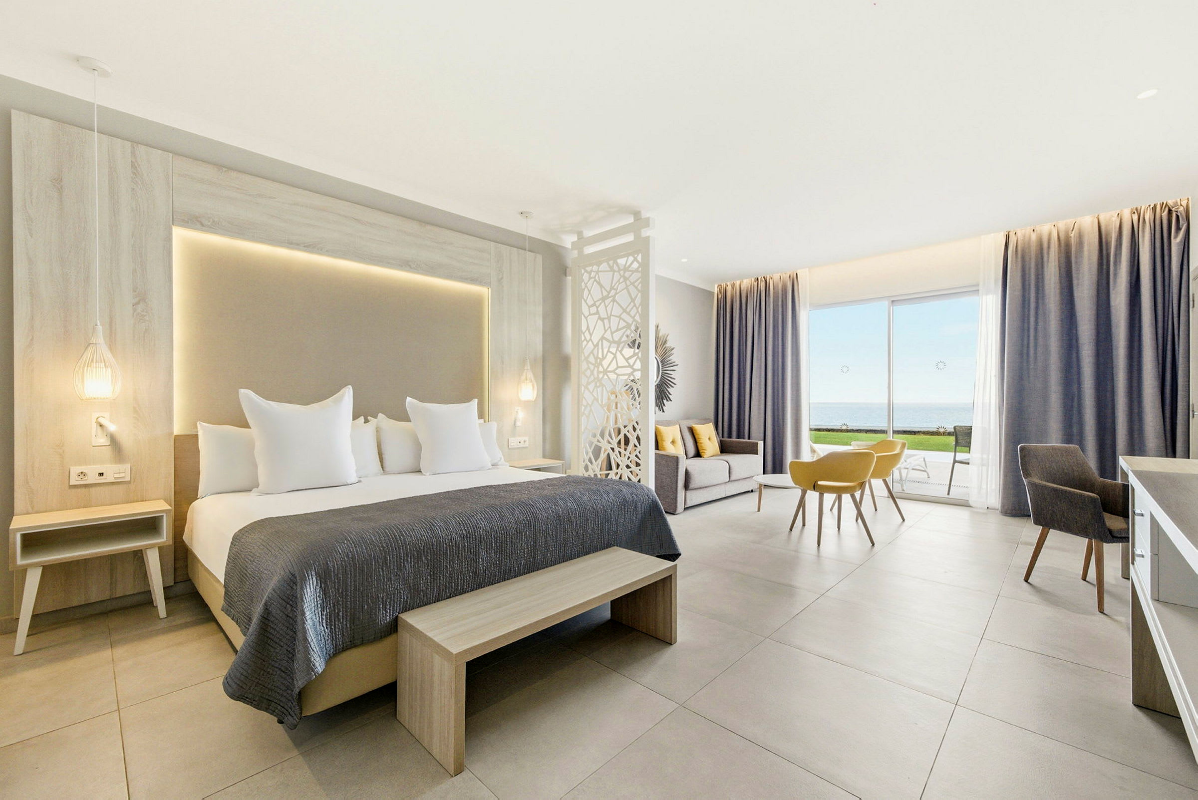 Et moderne hotelværelse med dobbeltseng, siddeområde og udsigt over havet gennem store glasdøre. (AI-genereret tekst)