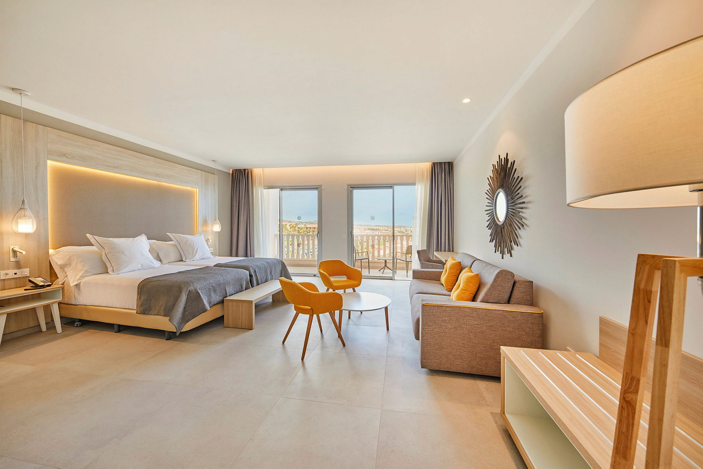 Et moderne hotelværelse med dobbeltseng, siddeområde med orange stole, en sofa og en balkon med udsigt til havet. (AI-genereret tekst)