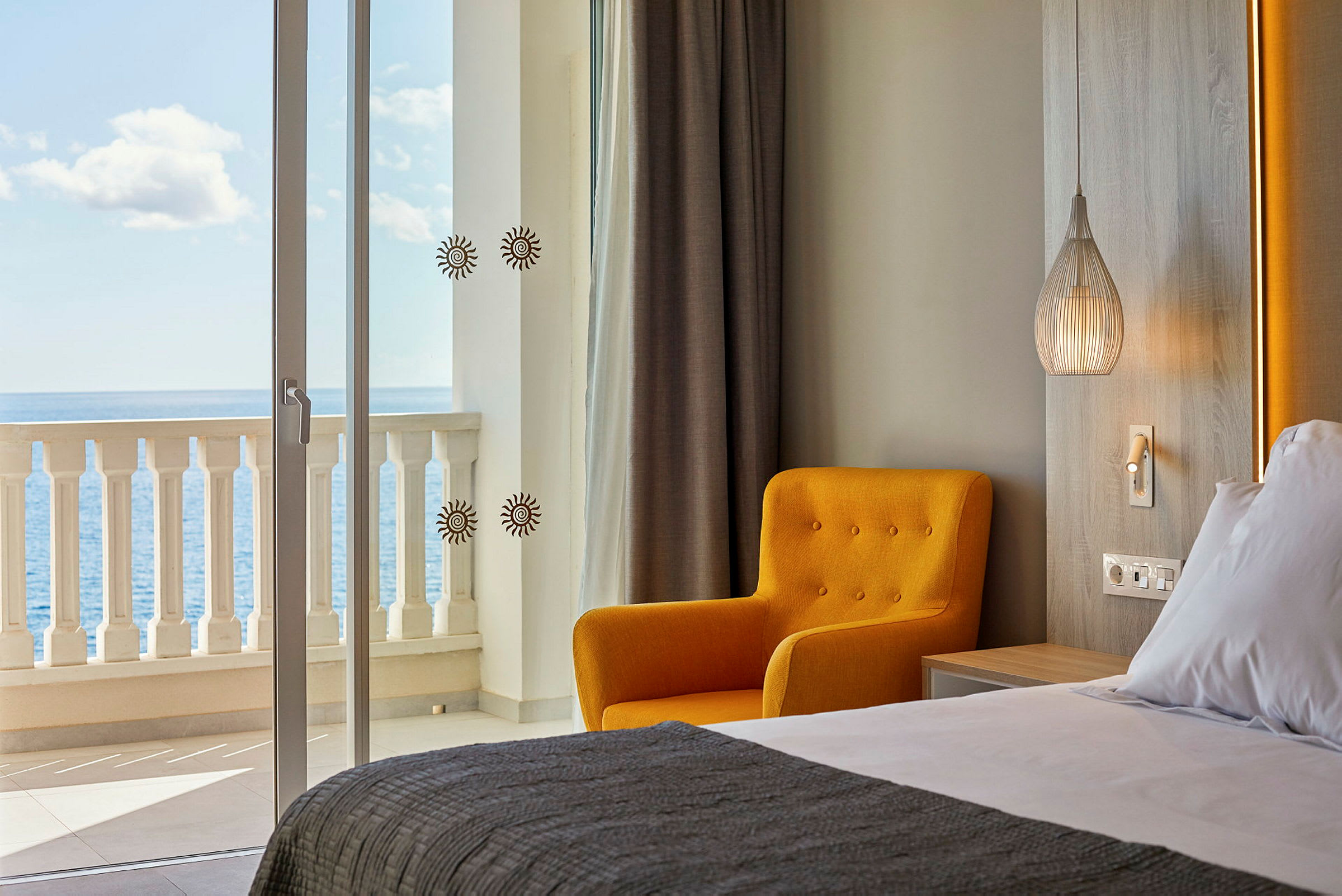 Et hotelværelse med havudsigt. I forgrunden ses en redt seng, sammen med en gul stol og en moderne lampe. (AI-genereret tekst)