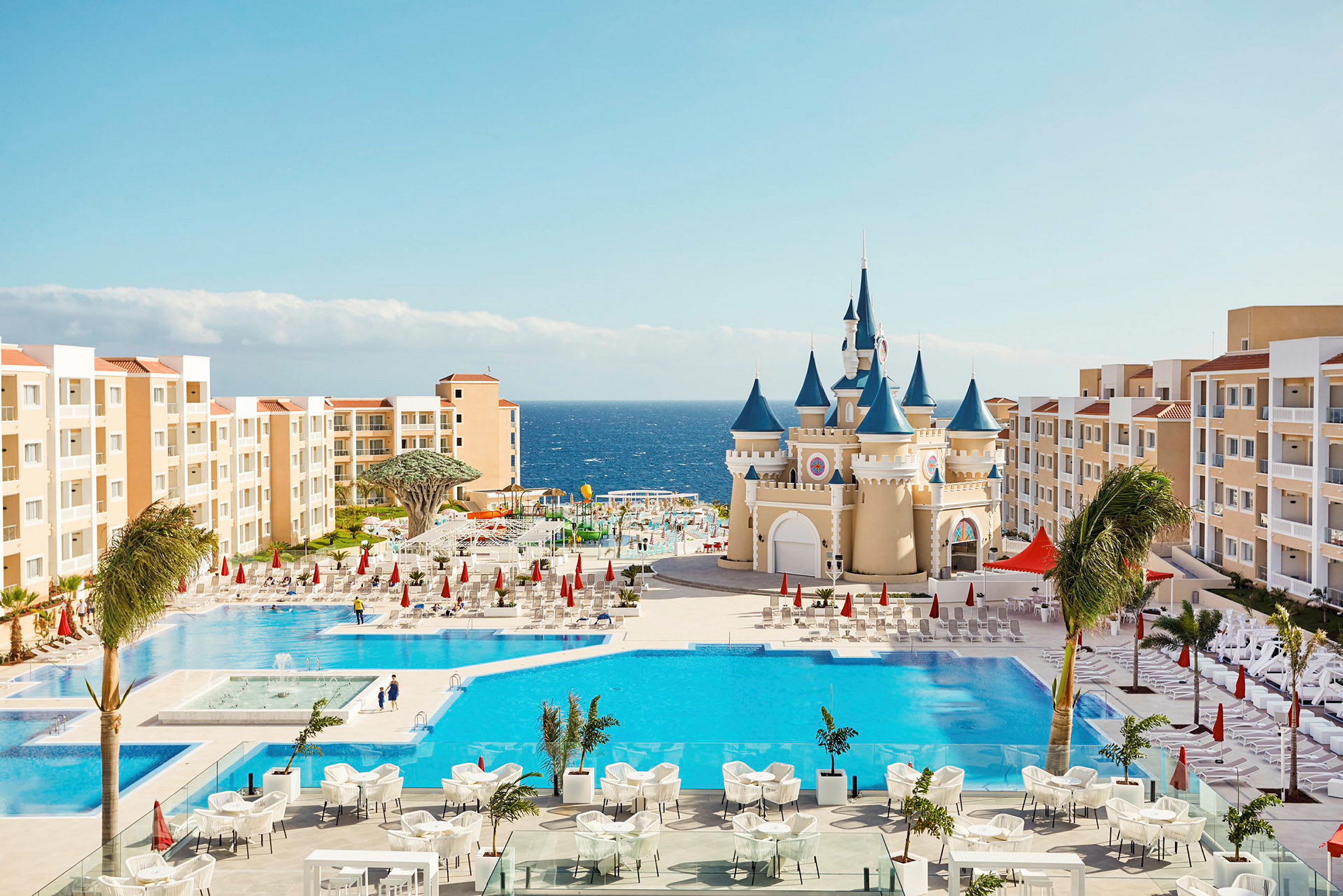 Et feriested med swimmingpools, en bygning der ligner et eventyrslot og omkringliggende hotelbygninger nær havet. (AI-genereret tekst)
