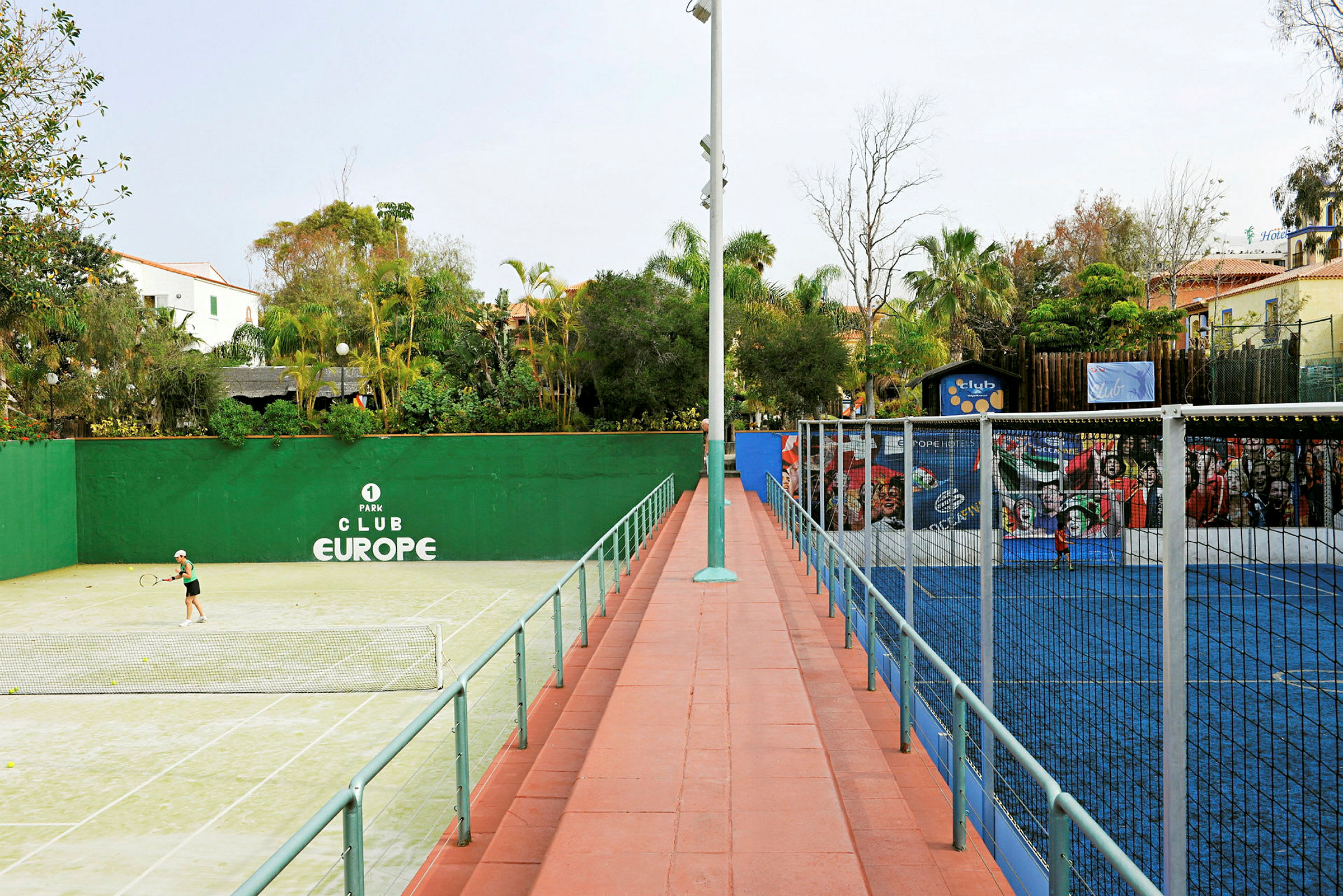 En grøn tennisbane til venstre og en blå padelbane til højre, omgivet af træer og bygninger. (AI-genereret tekst)